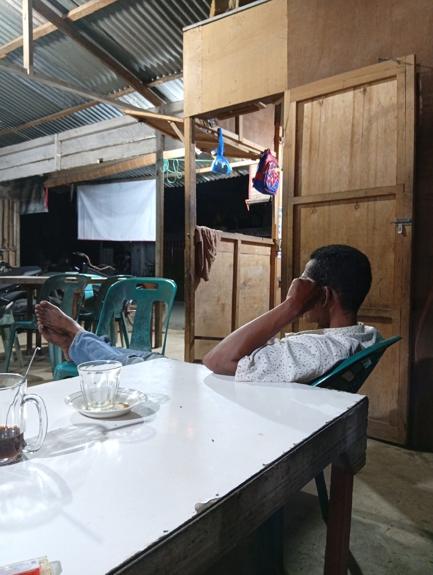 A man sits with his legs extended, resting on a green plastic chair in an outdoor seating area. He is wearing a white short-sleeved shirt with a repeating pattern of dark triangles and a dark blue-grey denim. His left foot is bare. In front of him is a white table with a glass mug containing a dark liquid, likely coffee, and a small empty glass on a saucer. The background shows a corrugated metal roof supported by wooden beams. There are several other green plastic chairs and tables visible, some with motorcycles parked nearby. A white banner hangs in the background. A wooden structure with a door is to the right. The scene appears to be a casual eatery or rest stop. The man is likely in Lhokseumawe, Indonesia, given the context of the request.