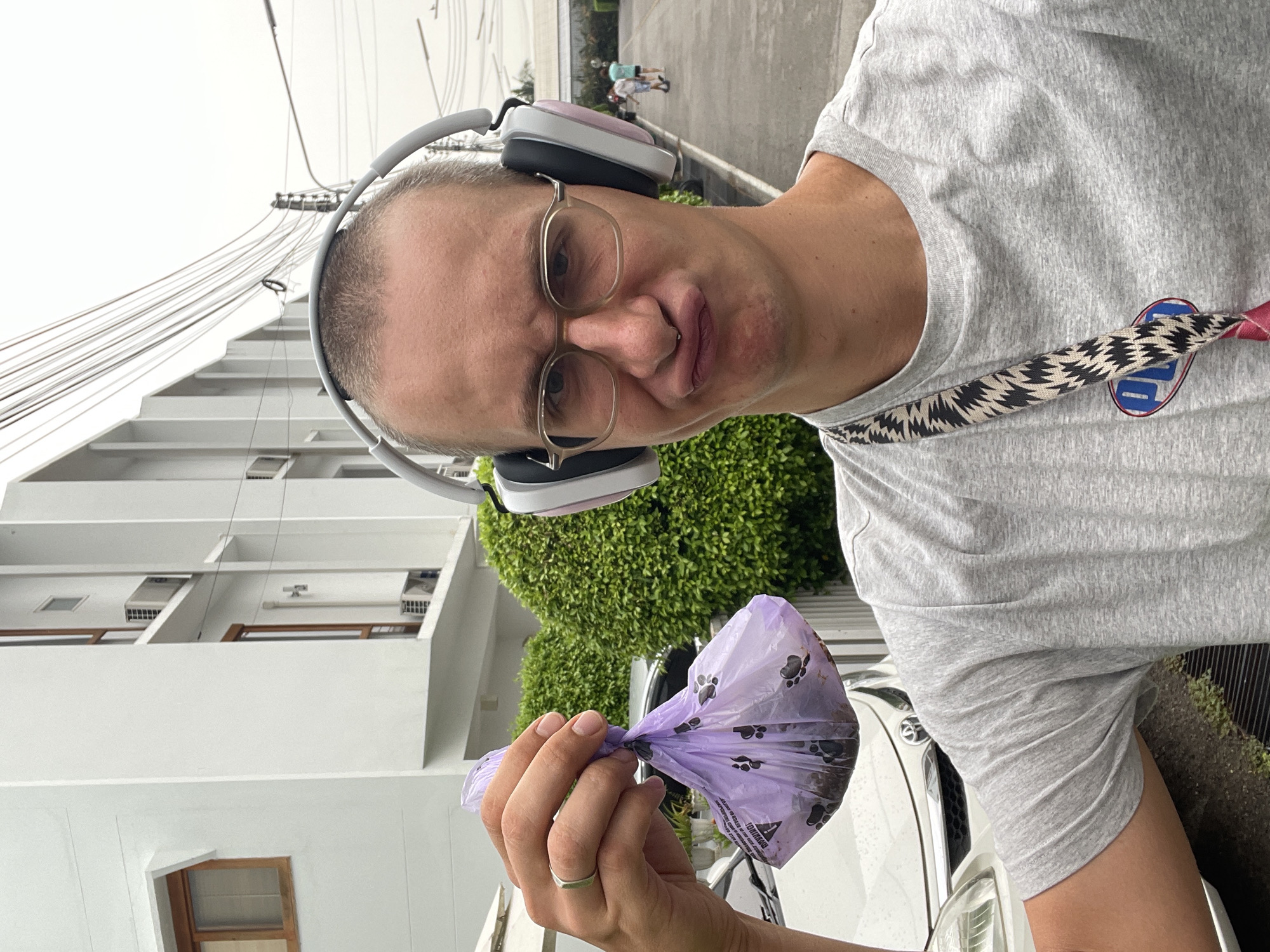 A man wearing headphones and glasses is holding a purple dog waste bag in his left hand. He is outside in Nong Pa Khrang, Thailand; a white building is visible in the background, as are power lines. The man is wearing a grey t-shirt and has a lanyard around his neck. A white car is partially visible in the background. Two people are further away, walking on the sidewalk.