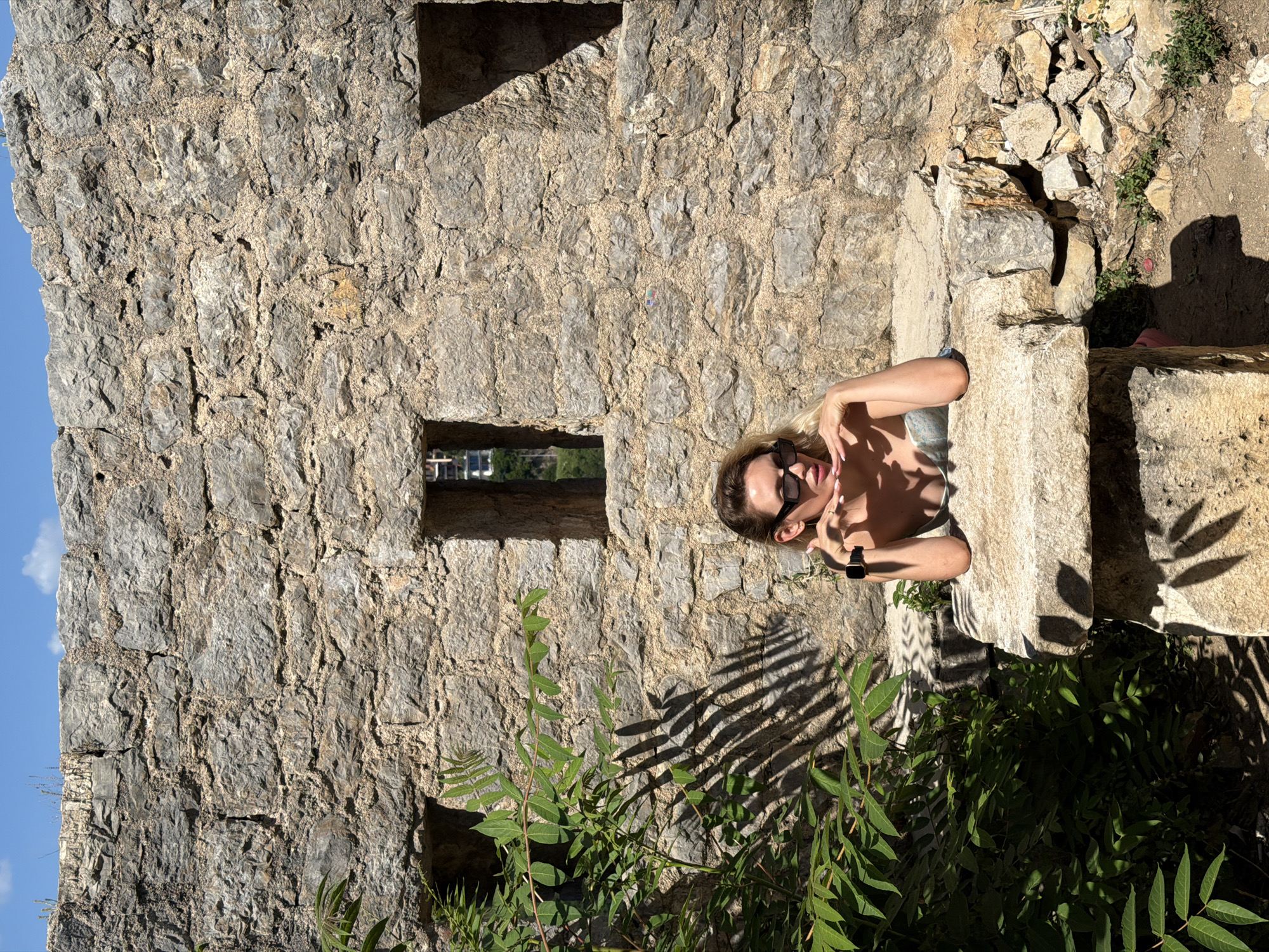 The image presents a sunlit scene in Budva, Montenegro, featuring a blonde female subject posing in front of an ancient, weathered stone wall. The wall is constructed from large, rough-hewn, irregular gray and brown stones bound with lighter-colored mortar. Three rectangular openings, or embrasures, are visible in the wall: one directly behind the subject's head, one to her left partially obscured by foliage, and one to her right. Through the openings, glimpses of distant buildings and green terrain are visible beneath a clear blue sky dotted with white clouds.
The female subject wears sunglasses and a light-colored, possibly strapless, top. She leans on a substantial, flat stone block extending from the wall, resting her forearms on it. Her right hand supports her chin, while her left hand is positioned near her mouth. A dark smartwatch is visible on her left wrist. She looks directly at the viewer.
To the left of the frame, a lush green plant with numerous leaves casts distinct, elongated shadows onto the stone wall and the ground below, indicative of direct overhead sunlight. The ground in the foreground and to the right consists of dry soil mixed with small stones and sparse vegetation.