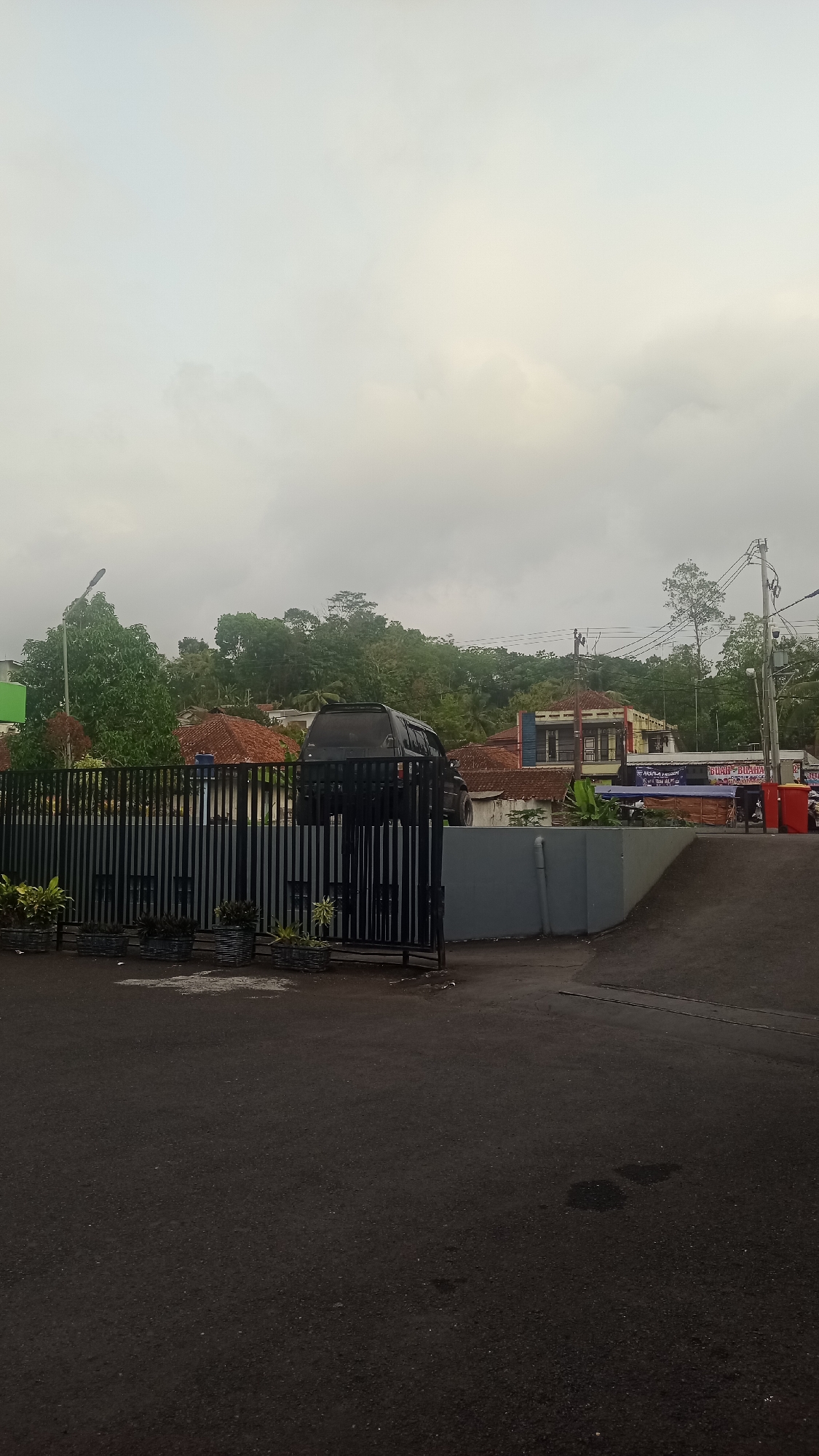 The image depicts a street scene in Karyamandala, Indonesia, under an overcast sky. In the foreground, a dark asphalt road occupies the lower half of the frame, showing some lighter patches. A black metal fence runs horizontally across the middle ground, with some planters containing green foliage placed in front of it. Behind the fence, a dark-colored SUV is parked. Beyond the fence, there are several buildings with red-tiled roofs, interspersed with lush green trees and vegetation covering the hilly landscape. Power lines and poles are visible against the sky. To the right, a blue and white banner is displayed, and a red trash bin is visible. A few people are seen near the banner and trash bin, indicating a public area. The overall impression is a rural or semi-urban setting.