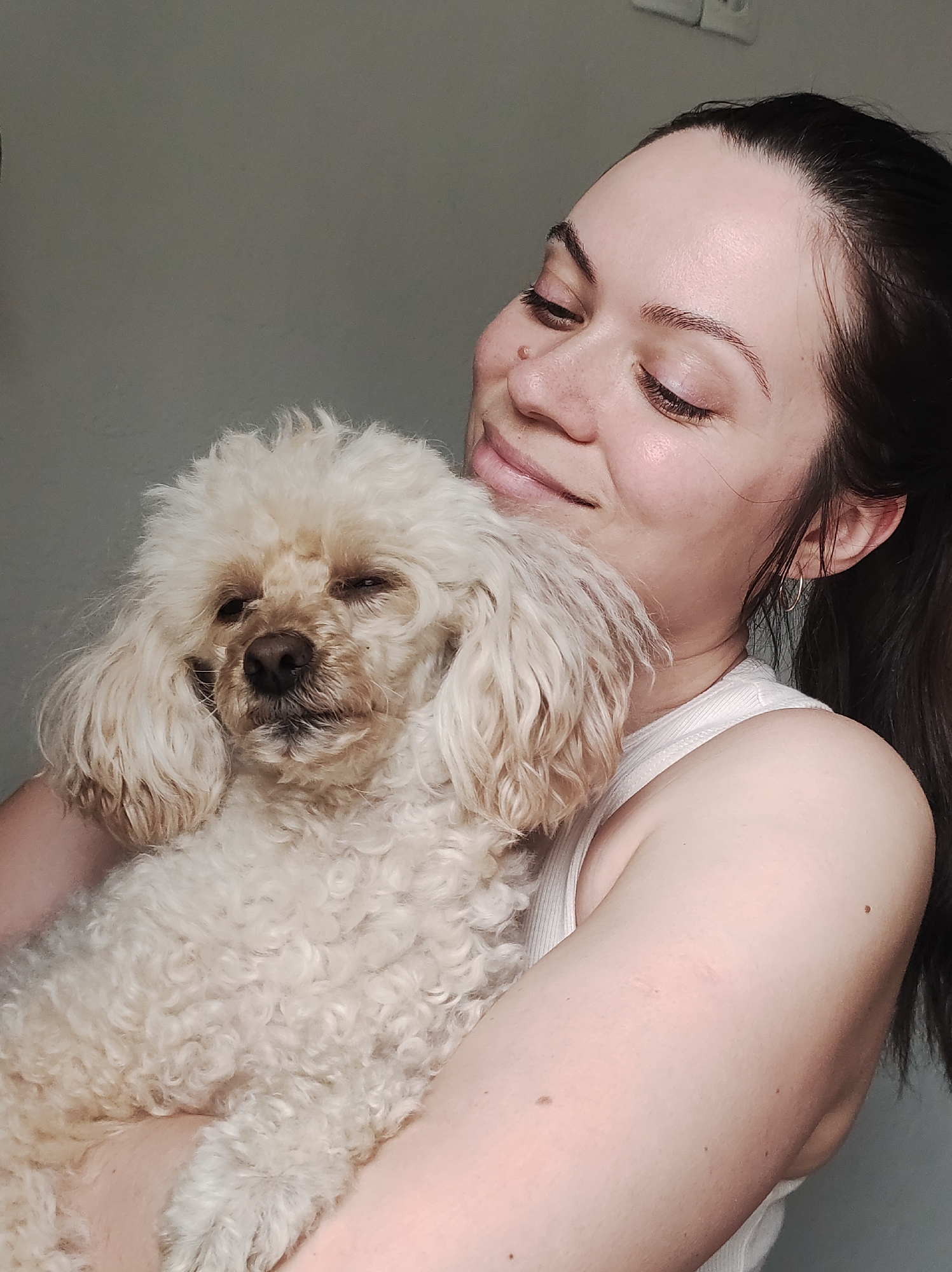A medium shot depicts a woman holding a small, cream-colored poodle. The woman has long, dark hair pulled back into a ponytail and is wearing a white tank top. She is looking down at the dog with a gentle smile. The dog appears sleepy, with its eyes partially closed. The background is a plain, light gray wall. No information available about location in Chaikovsky, Russia.