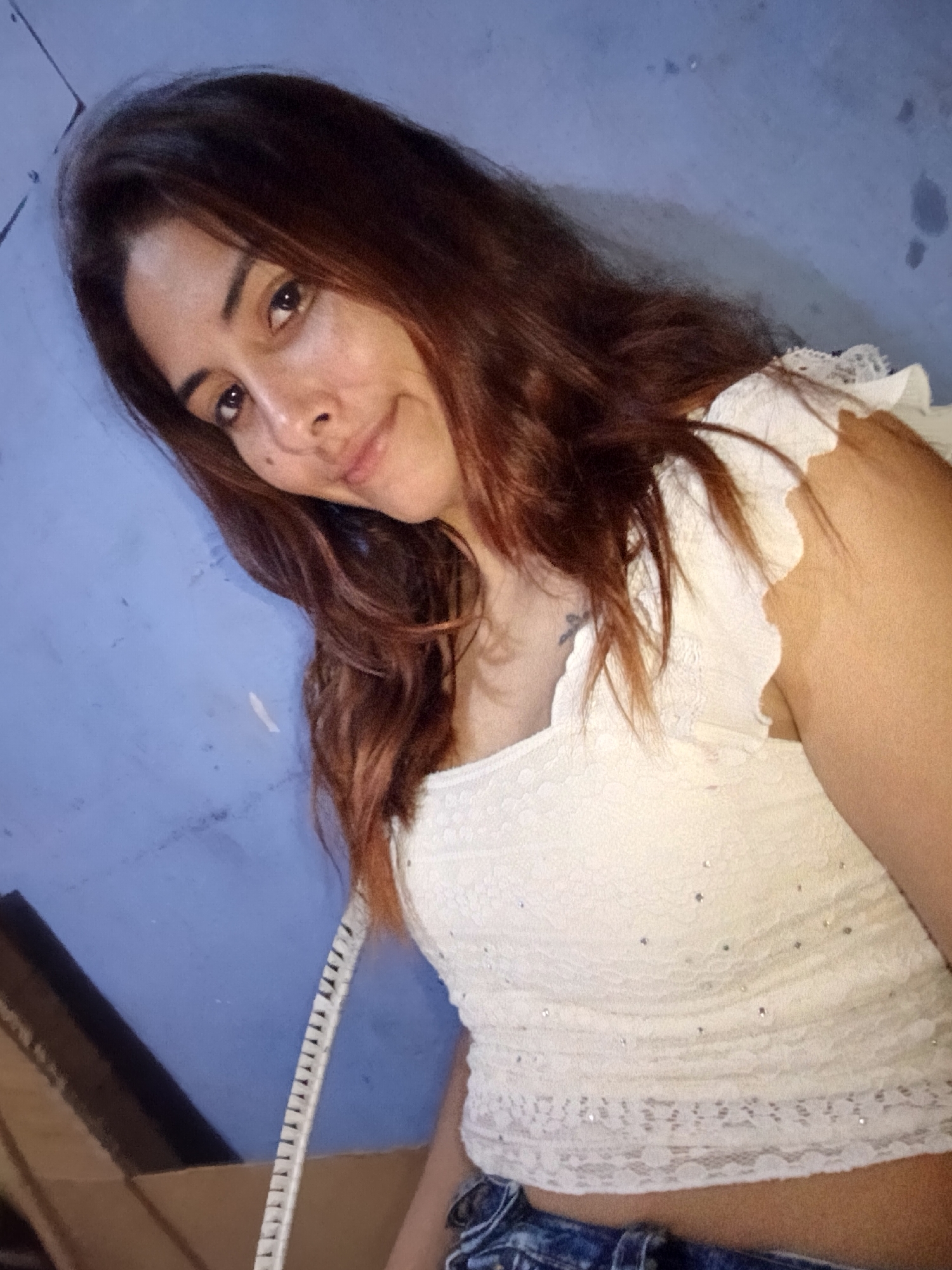 A young woman with long, auburn hair is leaning forward and looking at the camera. She is wearing a white, lace-trimmed crop top adorned with small, sparkling rhinestones, and blue denim shorts. A portion of a blue wall is visible behind her, with some smudges and marks on its surface. To the left of the woman, a cardboard box is partially visible. The image is a close-up, selfie-style shot, with the woman's face taking up a significant portion of the frame. The lighting suggests an indoor setting. The location in the city of Juan Griego, Venezuela, is not evident from the visual information in the image.