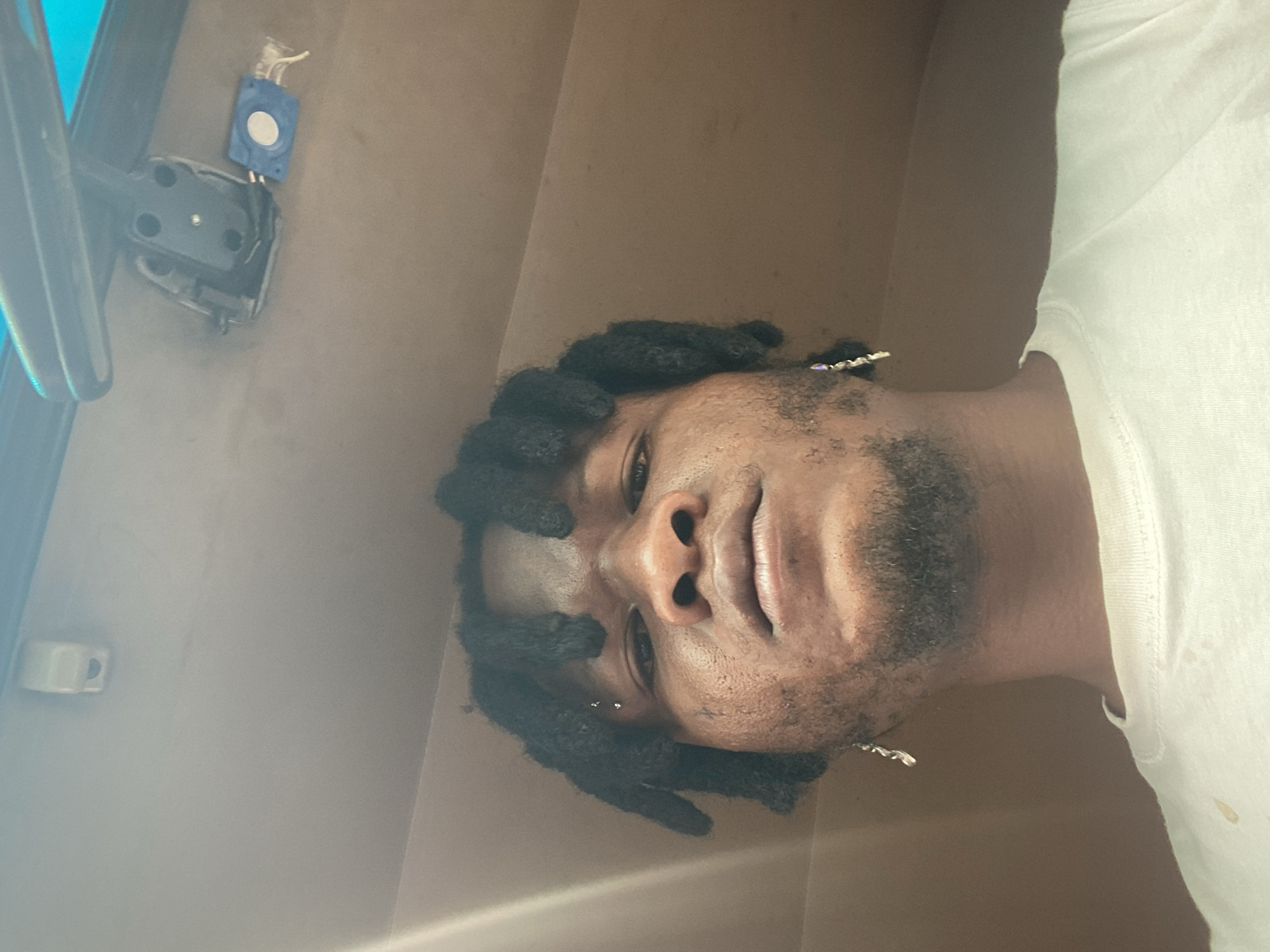A low-angle, close-up selfie is taken from within a vehicle, looking up at a Black male. He has dreadlocks, a small beard, and is wearing a white t-shirt and earrings. The ceiling of the vehicle is visible above him, with a rearview mirror and other car interior elements. There is a faint light source to the left of the frame, casting a glow on the subject's face and hair. The background of the image is a neutral, brown interior of the vehicle. No specific location within Jos, Nigeria is identifiable from the image.