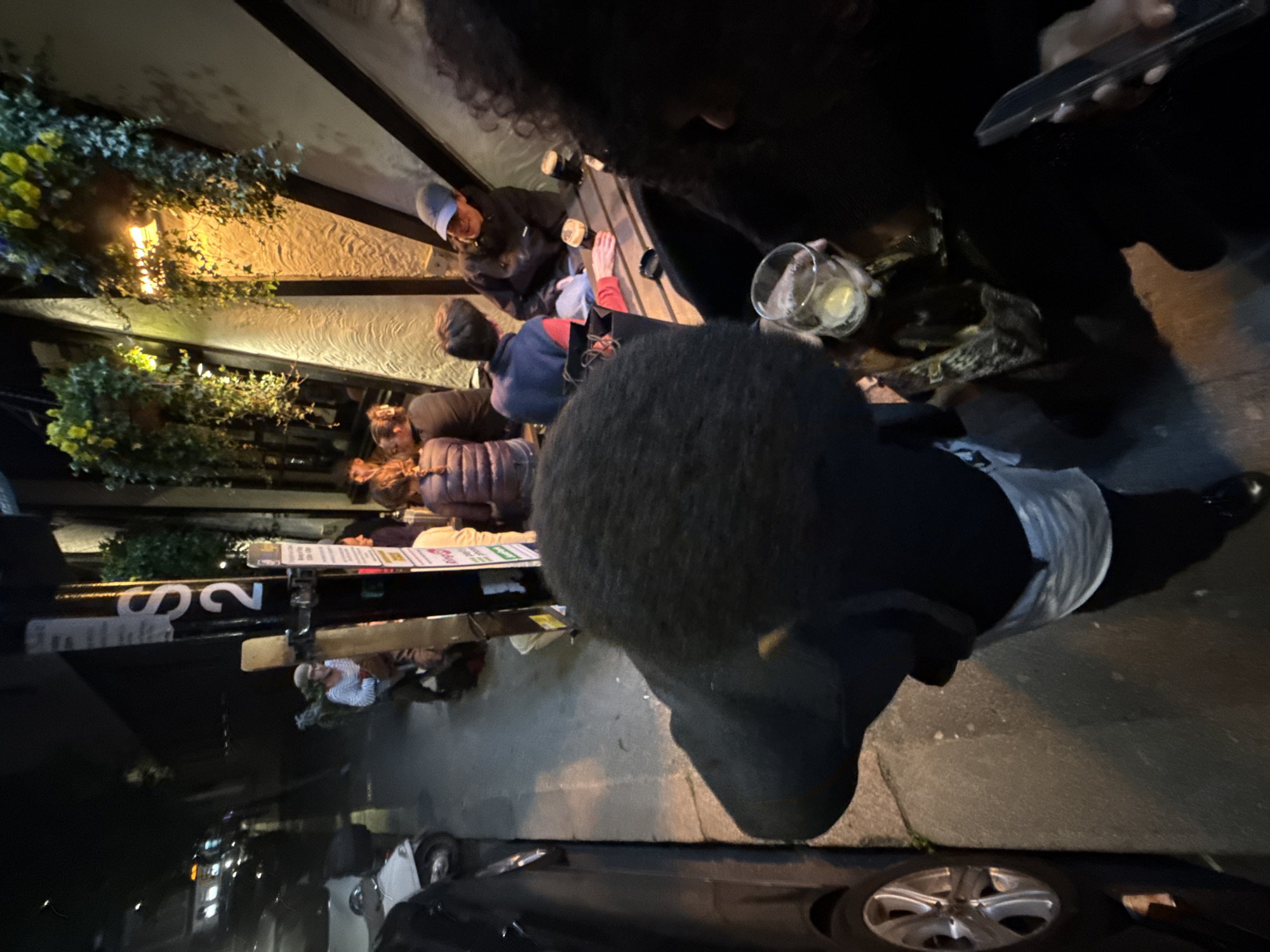 The image captures an outdoor scene on a London street at night. At least six people sit at a table outside a building, engaging in conversation and drinking. Hanging floral arrangements and string lights provide illumination. A street sign with the number "22" is visible in the background. A person, presumably taking the photograph, is holding a cell phone and has a drink in a glass. Parts of a black car are also visible, positioned on the street.