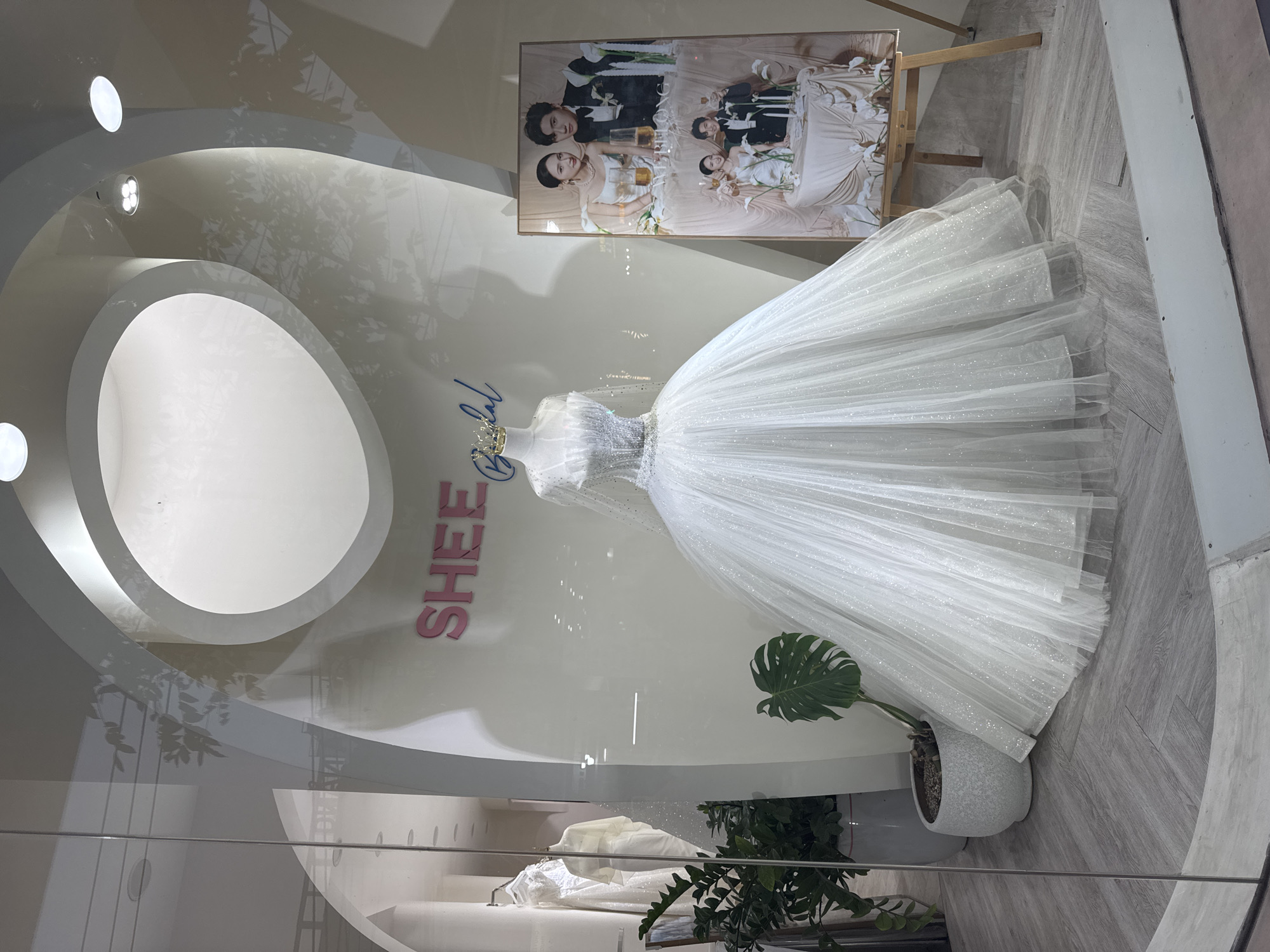 This appears to be a photograph of a wedding dress displayed in a shop window. The dress is a white, layered gown, possibly made of tulle or a similar fabric. It is displayed on a mannequin. Behind the mannequin, there is a sign with the word "SHEE" in pink letters, and a wedding photograph is propped up on an easel. A potted plant is present near the base of the mannequin. The window display features a unique architectural design with circular and curved white surfaces. The shop may be located in Đà Nẵng, Vietnam, but there is no irrefutable evidence in the image to confirm the location.