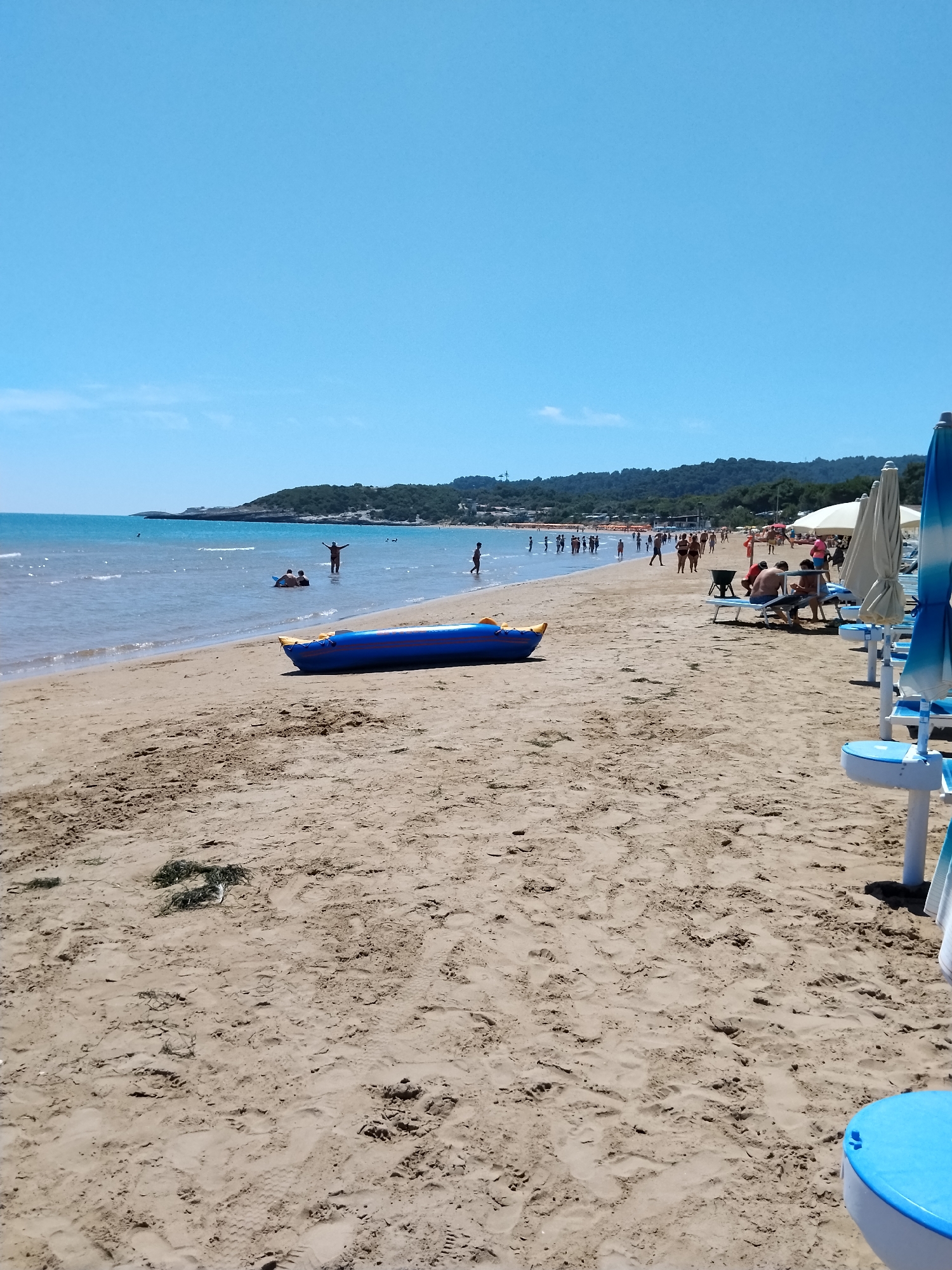 This image depicts a sunny day at a beach in Peschici, Italy. The foreground is dominated by sandy beach with footprints and some dried seaweed. In the midground, a blue inflatable banana boat rests on the sand. The sea is calm and light blue, with a few people visible in the water, some of whom appear to be on or near inflatable objects. Further down the beach, several rows of beach chairs and umbrellas are set up. A crowd of people can be seen walking and relaxing on the beach and near the water. In the background, a lush green, tree-covered hillside forms the coastline, with a clear blue sky above.