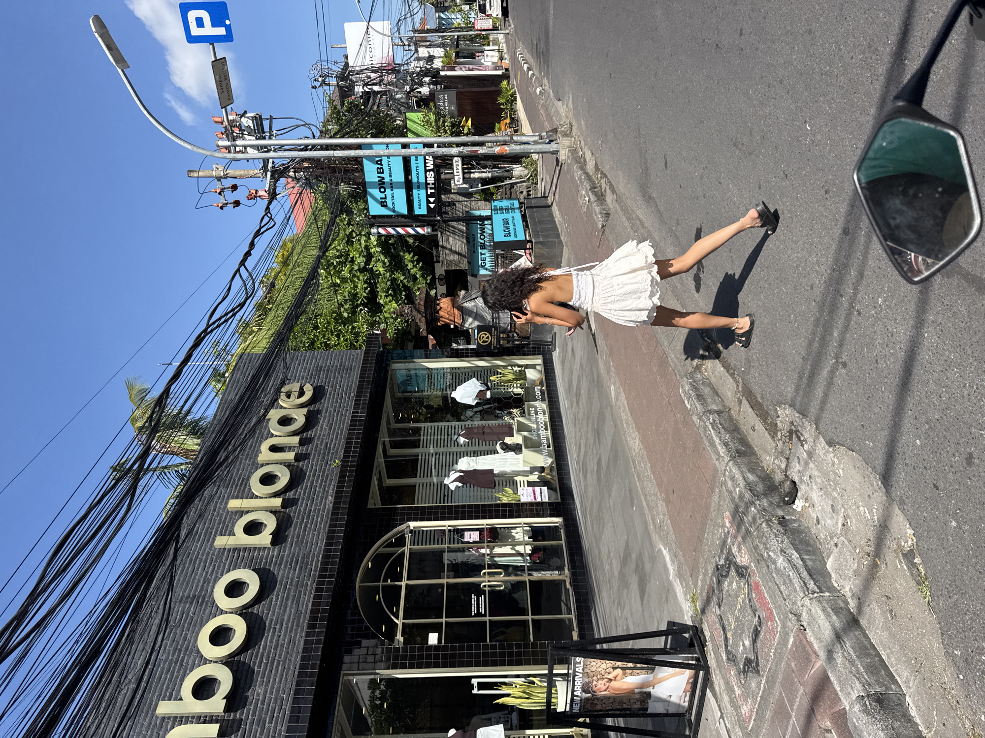 The image depicts a street scene in North Kuta, Indonesia. On the left, a shop with the name "Bamboo Blonde" is visible. A woman in a white dress walks along the sidewalk. Above the shop are tangled power lines. Adjacent to the shop are signs advertising "BLOW BAR", "BEAUTY", "BLOWOUTS", "BEAUTY", etc. A "P" parking sign is visible on a pole near the sidewalk. The street is paved, and a motorcycle mirror is visible in the top right.