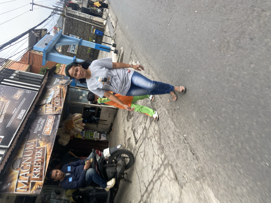 The scene is a street in Kecamatan Cimahi Selatan, Indonesia. A woman in a grey t-shirt and blue jeans is walking down the middle of the street with a young child in a green and orange tracksuit. The child is slightly behind the woman. The woman is looking straight ahead, and the child is looking to the left. The woman is wearing sandals, and the child is wearing white sneakers. The woman is walking on the left side of the street, and the child is walking on the right side of the street. There is a motorcycle parked on the right side of the street, and a man sitting on it. The man is wearing a dark blue shirt and black pants. He is looking straight ahead. There is a blue archway structure on the right side of the street. There are some buildings in the background, and there are power lines running across the street. The road is paved and has a few cracks. The sidewalk on the left side of the street is made of cobblestones. The sidewalk on the right side of the street is made of concrete. There are a few trees and bushes on the left side of the street. There are some trash cans on the right side of the street. There are some signs and banners on the left side of the street. The scene is taken in the daytime. The weather is clear.