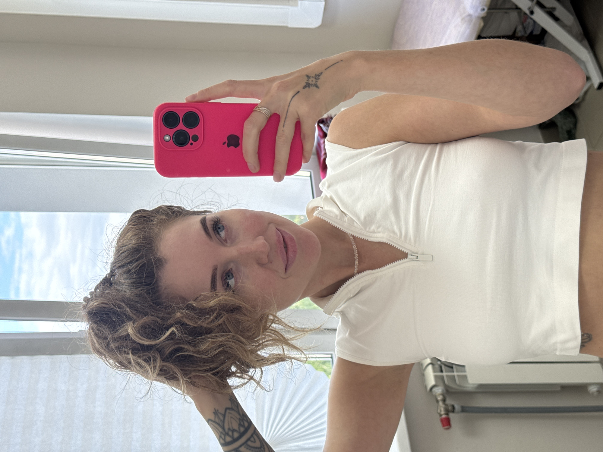 A young woman with curly brown hair, blue eyes, and a light complexion takes a mirror selfie indoors. She is captured from the waist up, smiling softly and looking directly into the camera's reflection. She wears a white, short-sleeved top with a quarter-zip neckline and a delicate silver chain necklace.
Her right hand, adorned with a silver multi-band ring on the ring finger and a small star/compass rose tattoo, holds a bright pink smartphone. The phone, identifiable as an iPhone by its triple camera array and visible Apple logo, is positioned for the selfie. A larger, intricate mandala-style tattoo is visible on her left forearm.
The background shows an indoor setting, likely a room in Chaikovsky, Russia. A window on the left is partially covered by white horizontal blinds, revealing a blue sky with some clouds and green foliage outside, indicating daytime. On the right side of the image, a white radiator with grey and red pipes is visible in the lower portion, and some indistinct furniture or shelving is in the upper background. The scene suggests a casual and personal moment captured in natural light.