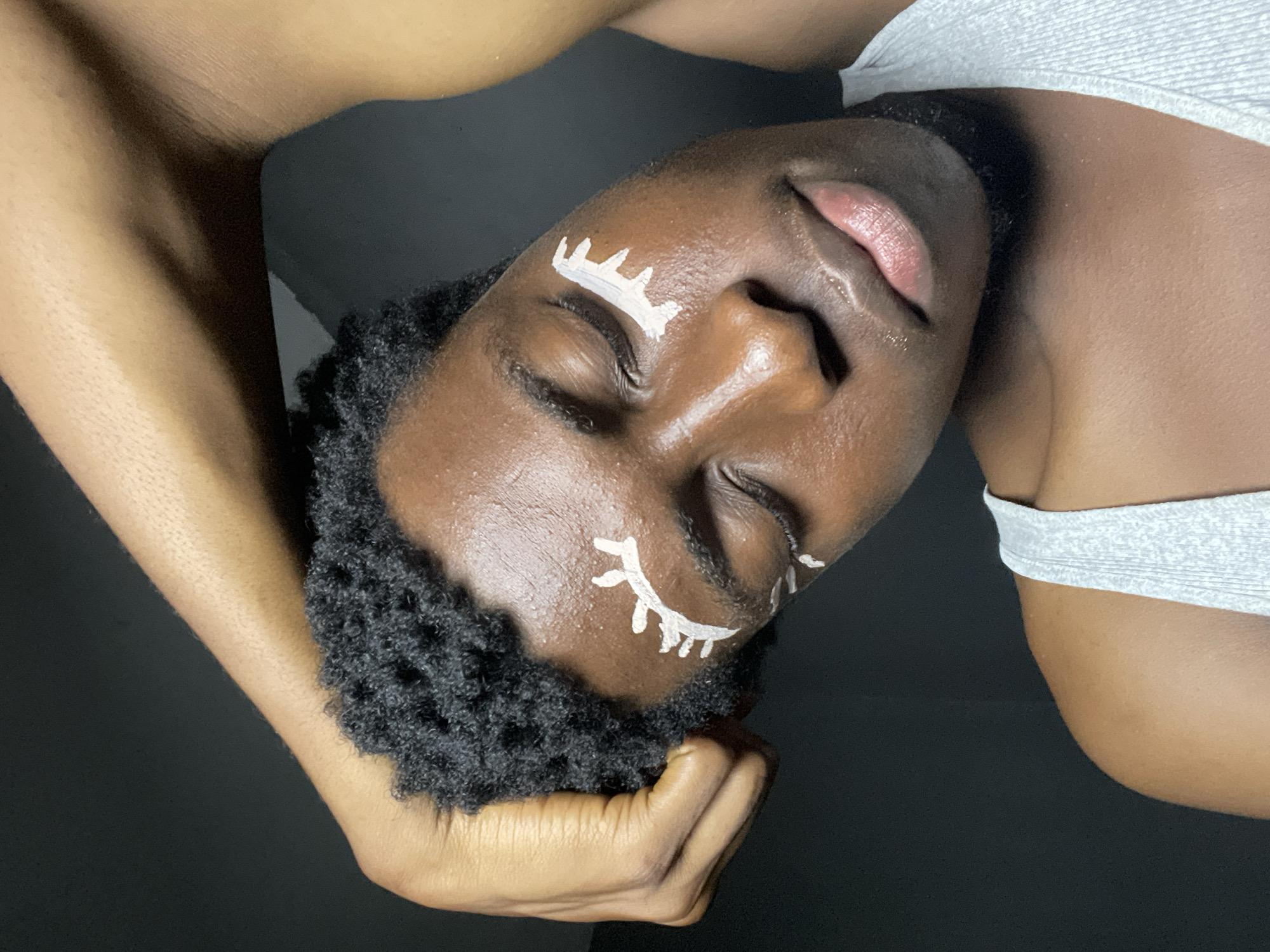 The image is a close-up shot of a young Black person with dark, curly hair and closed eyes. They are wearing a light gray tank top. White, artistic markings resembling stylized eyelashes are painted on their eyelids and under their eyes. The person's left arm is raised and bent, with their hand resting on their head, obscuring part of their hair and temple. The background is dark and out of focus, suggesting an indoor setting. There are no visible landmarks or contextual clues within the image to suggest a specific location in Nkpor, Nigeria.