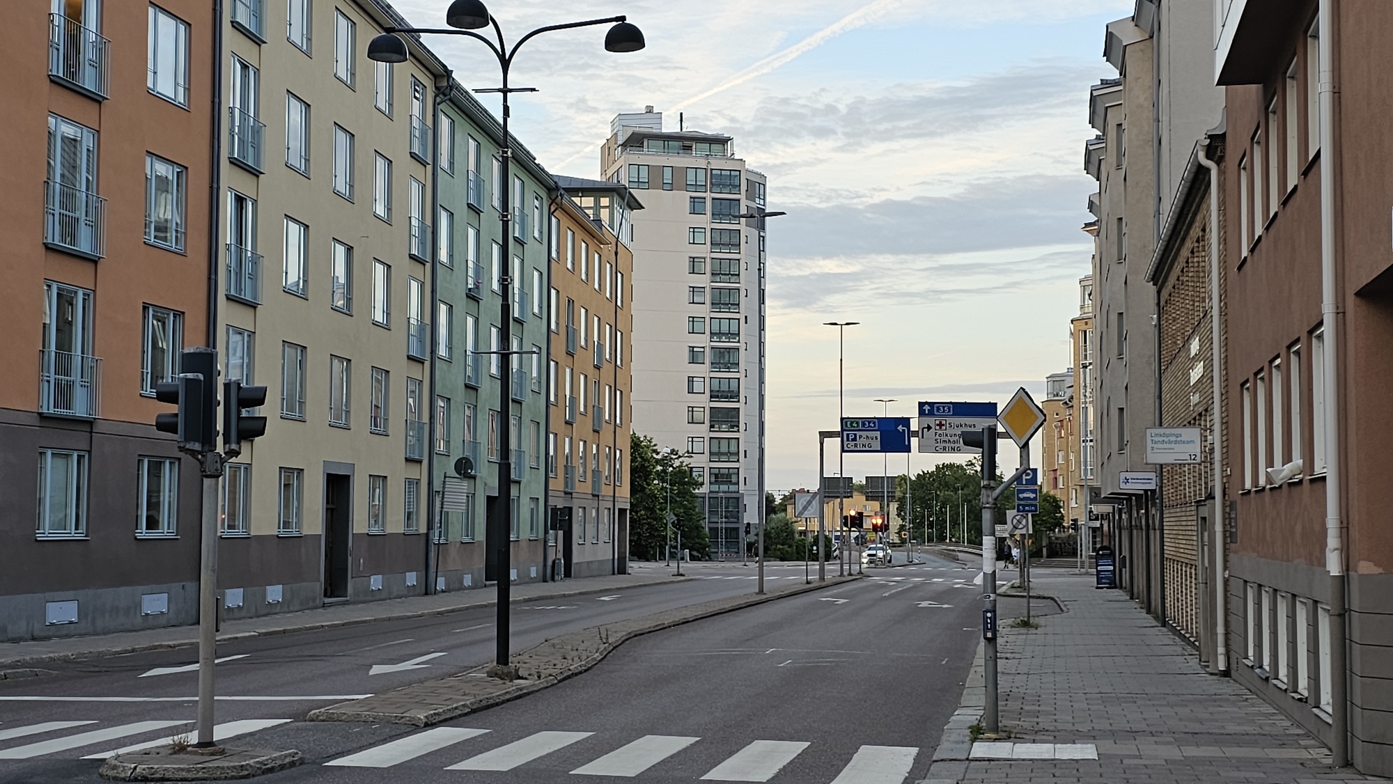 This is a wide-angle shot of a city street in Linköping, Sweden, likely in the late afternoon or early evening, given the soft, diffused light. The sky is a pale blue with wispy white clouds, suggesting fair weather. The scene is dominated by apartment buildings of varying heights and colors lining both sides of the street. On the left, buildings are painted in warm tones of orange, pale yellow, and green, featuring numerous windows and balconies. A tall, modern white building stands further down the street on the left. On the right, buildings are rendered in shades of beige and brown brick, with a more uniform architectural style.
The street itself is paved and features a pedestrian crosswalk in the foreground. Traffic lights and street lamps are visible, indicating an active urban environment. Several road signs are present, including directional signs for "E4 34", "P-hus", and "C-RING", as well as signs indicating "Sjukhus" (hospital), "Folkung", "Simhall" (swimming hall), and a sign for "Linköpings Tandvårdsteam" (Linköping Dental Team) with the number 12. A yellow diamond-shaped sign with a black border is also visible, likely a warning sign.
There are no people clearly visible in the immediate foreground, but a few figures can be seen further down the street on the right sidewalk. A car is also visible in the distance on the road, suggesting normal traffic flow. The overall impression is of a calm, everyday urban scene in a Swedish city.