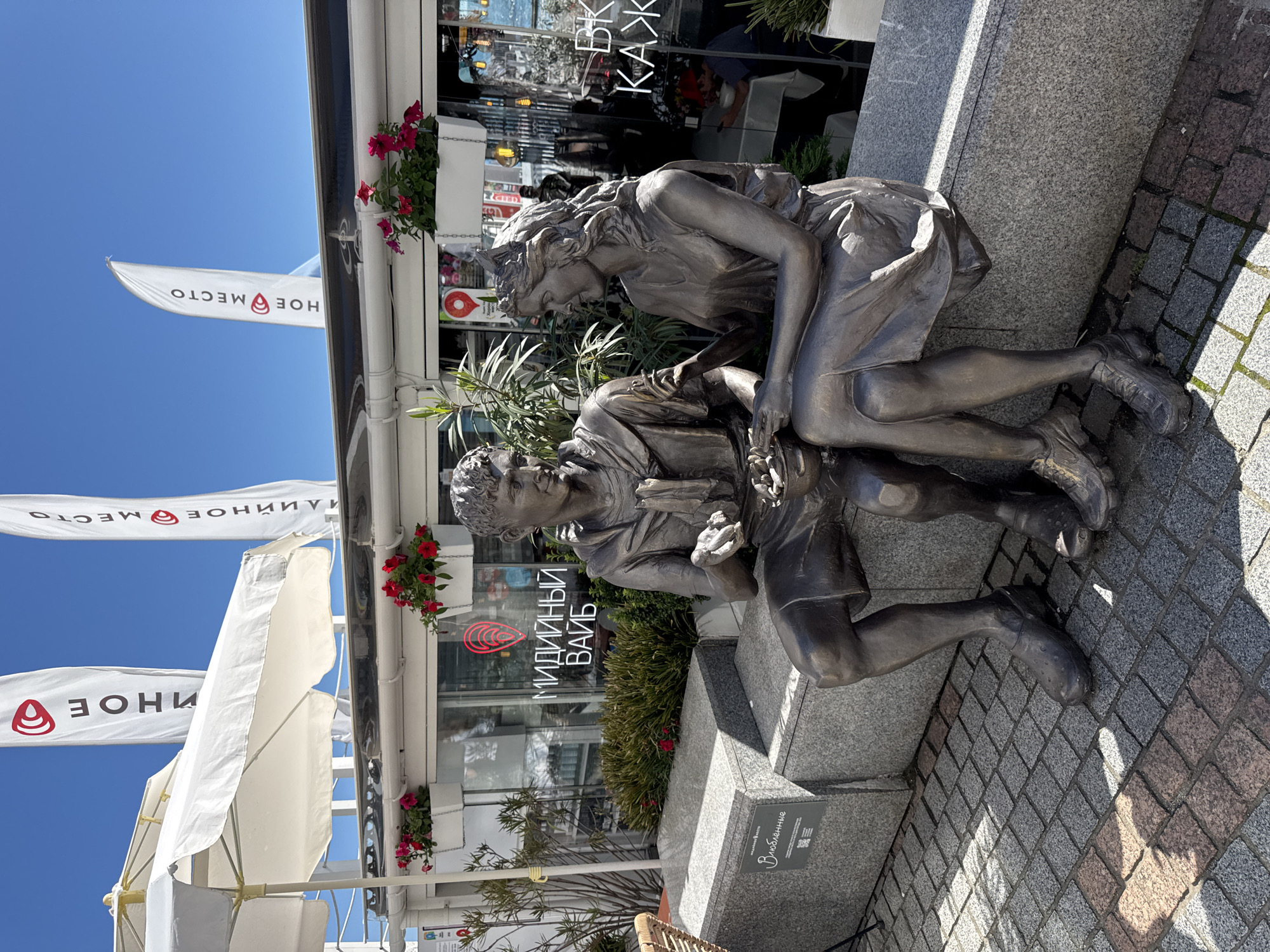 The image captures a bronze sculpture of a young couple on a sunny day in Sochi, Russia. The statue features a boy seated on a granite bench, looking down at something he holds delicately in his hands, possibly a small animal or object. A girl leans over him from the right, her gaze also fixed on the item, conveying a moment of shared curiosity or affection.
In the background, a modern building with large glass windows reflects the surroundings. Vibrant red petunias spill from planters adorning the building's facade. Several white flags flutter against a clear blue sky; visible text on them includes "МОРЕ МЕСТО" (Sea Place) and "ХОЕ МЕСТО" (possibly part of "Хорошее Место" - Good Place, or "Зонное Место" - Zoned Place). The building itself features signage such as "МИДИЙНЫЙ ВАЙБ" (Mussel Vibe), likely a restaurant, and partial text "ВКУС КАЖД" (Taste of Each). White outdoor umbrellas suggest a cafe or restaurant patio.
The statue is positioned on a granite base, beside which a sign reads "Влюбленные" (Vlyublennye), meaning "Lovers" or "In Love," along with a QR code. The ground is paved with multi-toned cobblestones, and sharp shadows indicate bright daylight and clear weather. The scene depicts a charming public art installation integrated into a lively commercial area, characteristic of a tourist-friendly promenade.