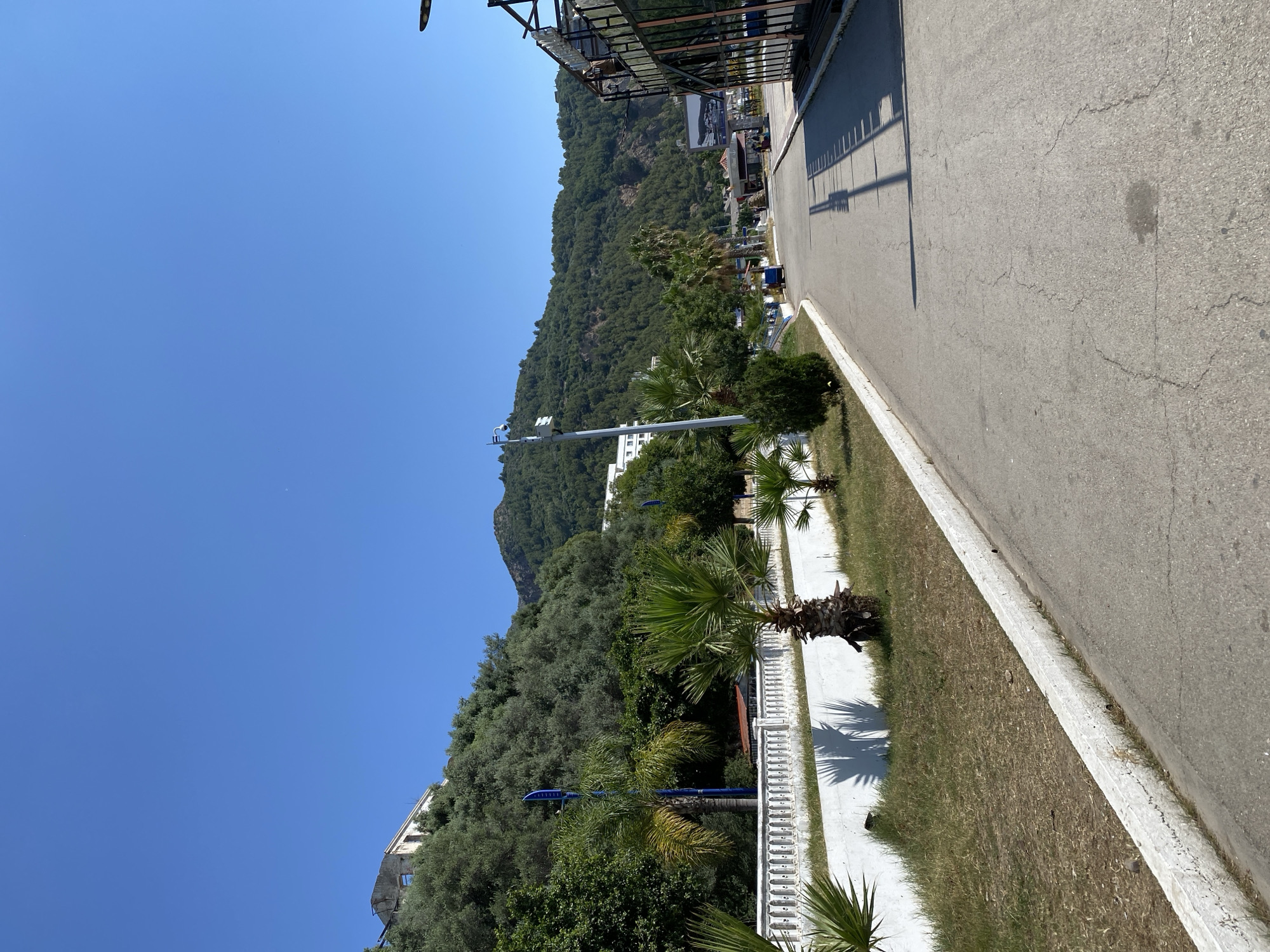 This media file captures a bright, sunny outdoor scene in Bejaia, Algeria, featuring a street running parallel to a lush, tree-covered mountain. The clear, vibrant blue sky, free of clouds, fills the upper left portion of the frame, suggesting excellent weather.
In the foreground and extending towards the mid-ground, a grey asphalt road is visible on the right, showing some surface cracks and imperfections. Long, sharp shadows, likely cast by unseen structures overhead, stretch across the road, indicating a time of day from mid-morning to early afternoon. Adjacent to the road, a white concrete curb borders a strip of dry, patchy grass. Beyond the grass, a white concrete sidewalk or paved area is present, intermittently lined with decorative white balustrades.
Several palm trees with prominent green fronds are interspersed along the sidewalk and grassy verge, casting distinct shadows onto the concrete. Among these trees, a few blue-colored lamp posts or decorative poles are also visible. A white pole with attached equipment, possibly a security camera or sensor, extends into the lower-left foreground, pointing towards the mountain.
The left side of the image is dominated by a densely vegetated mountainside, covered with a thick canopy of various green trees, including what appear to be olive trees. Towards the base of the mountain, a white building or complex is discernible. Further along the road in the mid-ground, additional buildings are visible, some featuring dark, possibly metal, canopies or overhead structures. One building has a large sign or billboard, though any text on it is illegible due to distance.
No people or specific activities are depicted in this static street view. The overall impression is one of a warm, bright day in a scenic Mediterranean coastal or mountainous region.