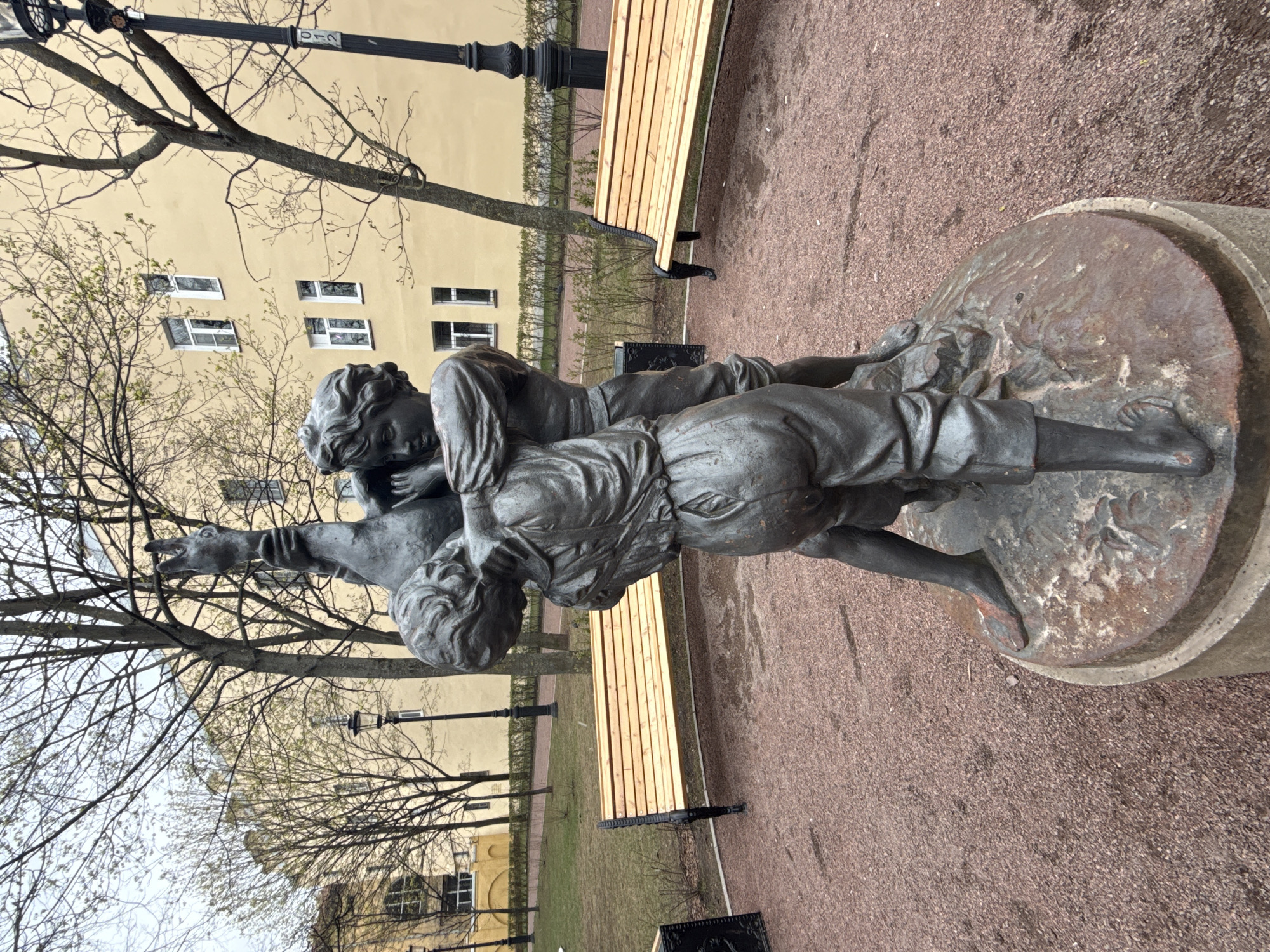 The media file presents a bronze statue located in a park or public square in Saint Petersburg, Russia. The statue depicts a young boy in a dynamic pose, carrying a lean greyhound dog. The boy, wearing what appears to be period attire including knee-length trousers, cradles the dog against his chest. The dog’s head is raised with its mouth slightly ajar, and its front paws rest on the boy's shoulder. The boy's right arm extends forward, suggesting a pointing or leading gesture, while his left arm supports the dog. The statue is mounted on a circular, multi-tiered base.
The setting includes a light yellow building in the background with multiple rectangular windows, some framed in white. The scene is dominated by deciduous trees with bare branches, some showing early signs of green foliage, indicating springtime. Park elements visible are a long wooden bench with dark metal supports and a dark, ornate lamppost. The ground surface is reddish-brown gravel. The lighting is soft and diffused, characteristic of an overcast day. Text "D 210" is visible on the lamppost.