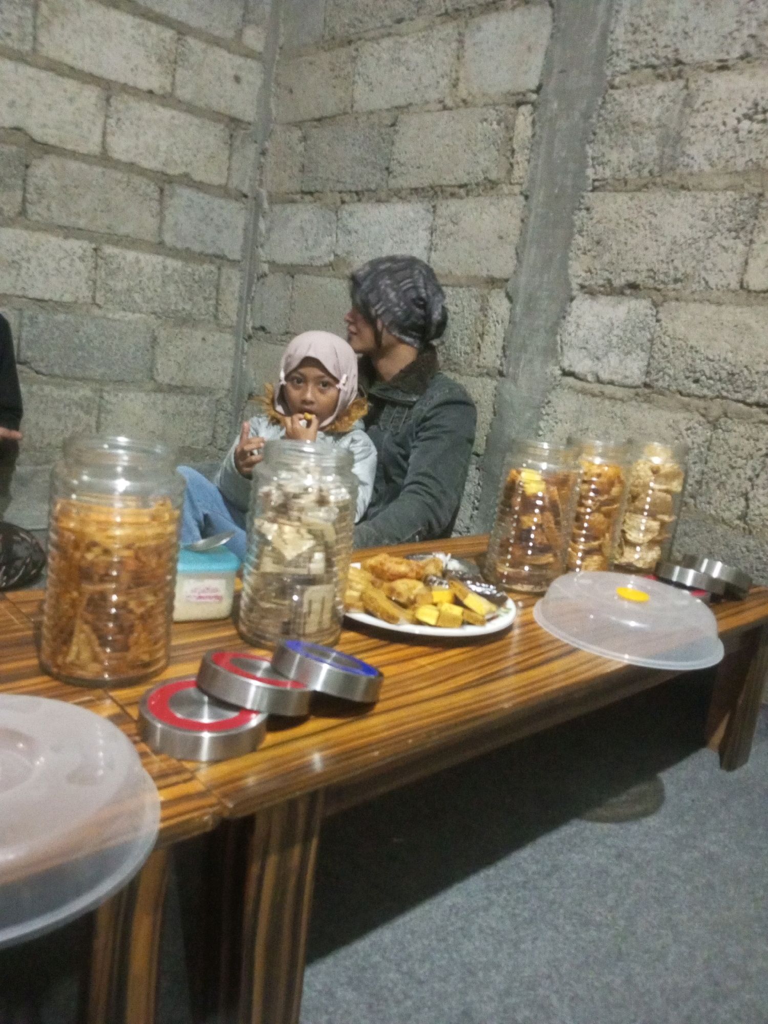 In Keseneng, Indonesia, a young child in a light pink hijab and a gray jacket is being held by an adult wearing a gray beanie and a dark jacket. They are sitting at a wooden table in front of a brick wall. On the table are several large glass jars filled with snacks, a plate of food, and a covered dish. Two metal rings with red and blue accents are in the foreground.