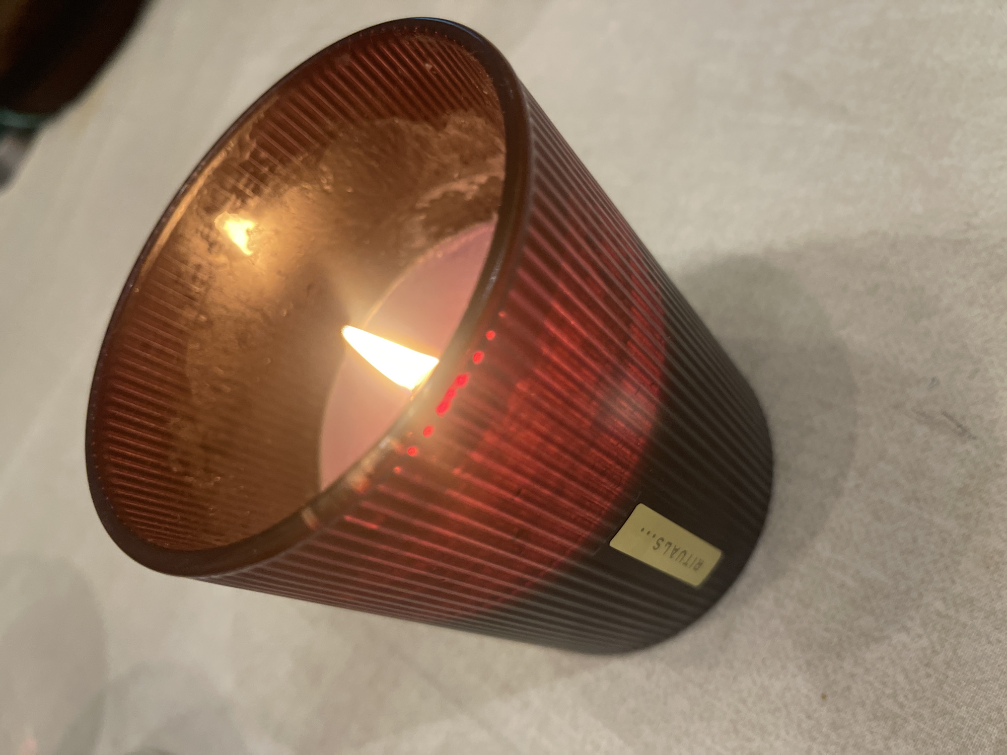 A close-up shot shows a lit candle in a textured glass holder, with a label that reads "RITUALS..." affixed to the side. The candle is placed on a white surface. The city and specific location are unknown.