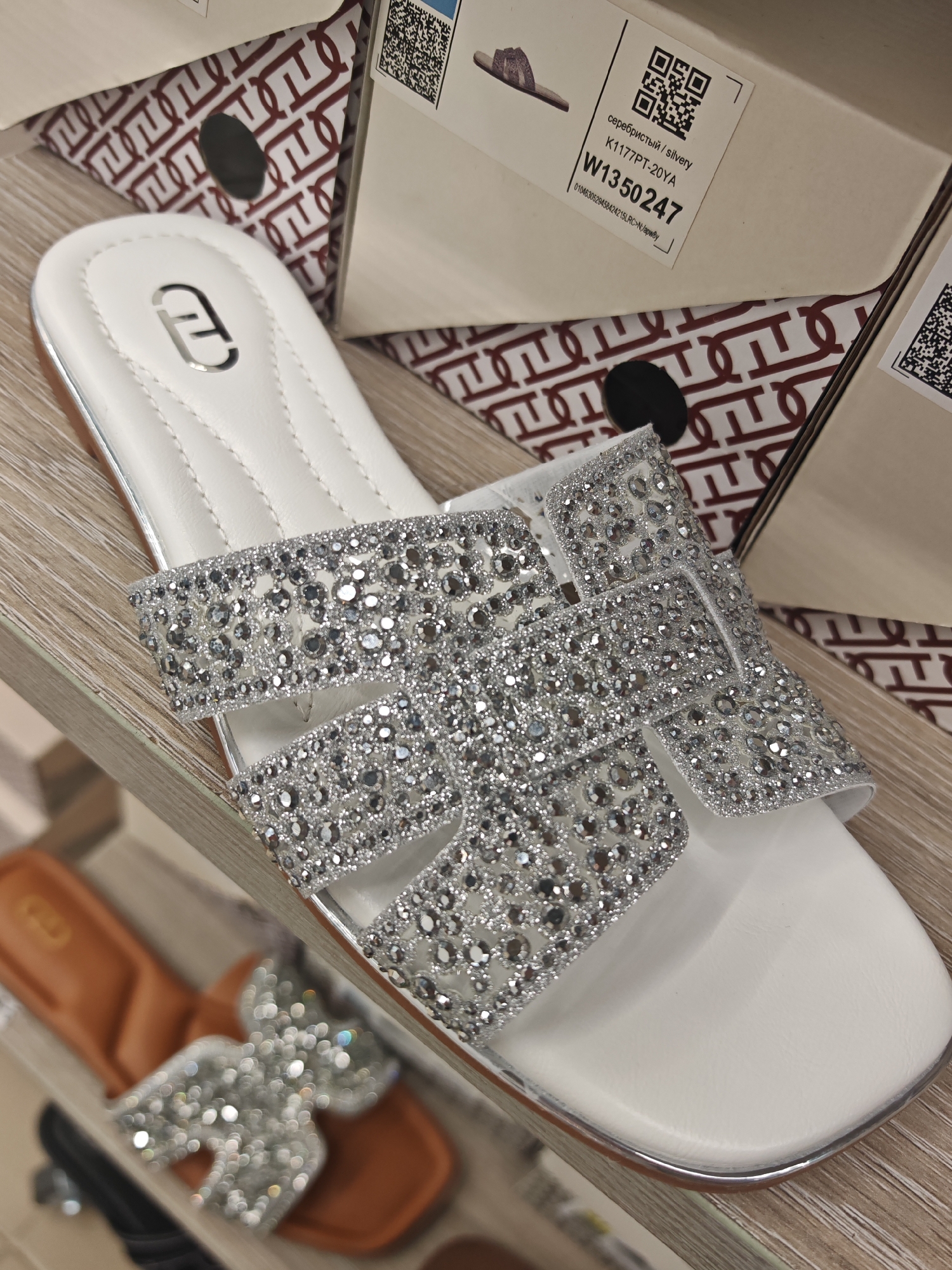 This media file shows a close-up of a pair of white slide sandals adorned with numerous sparkling rhinestones across the straps. The sandals are resting on a wooden shelf, and in the background, a shoe box with a repeating logo pattern and product information is visible. The scene appears to be a retail environment, likely a shoe store. The lighting is bright, suggesting an indoor setting. One of the cropped images reveals a label on the shoe box with the text "серебристый / silvery", "K1177PT-20YA", "W1350247", and a barcode. Another cropped image shows a second pair of brown slide sandals with a large, sparkly bow on the strap, also on display. The overall impression is one of a product display for fashionable footwear. The location context provided is Novy Urengoy, Russia.