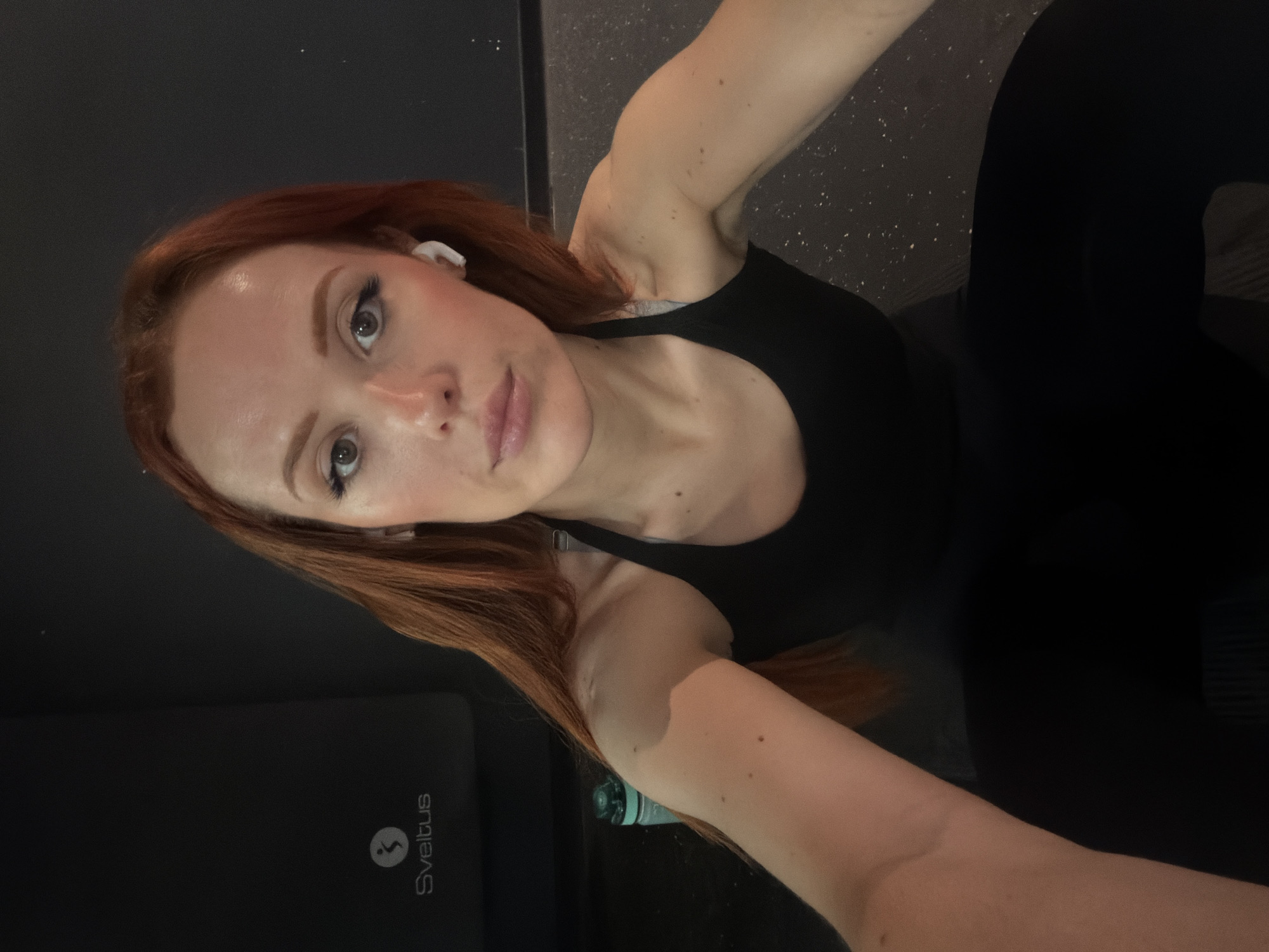 A woman with auburn hair is taking a selfie in what appears to be a gym setting. She is wearing a black tank top and black leggings. A white wireless earbud is visible in her left ear. A black exercise mat with the logo "Sveltus" is partially visible in the lower-left corner of the frame. A water bottle is also partially visible near the mat. The background consists of a dark wall and a floor with a speckled pattern. Based on the "Sveltus" branding, it is possible the image was taken in Paris, France, as Sveltus is a French company.
