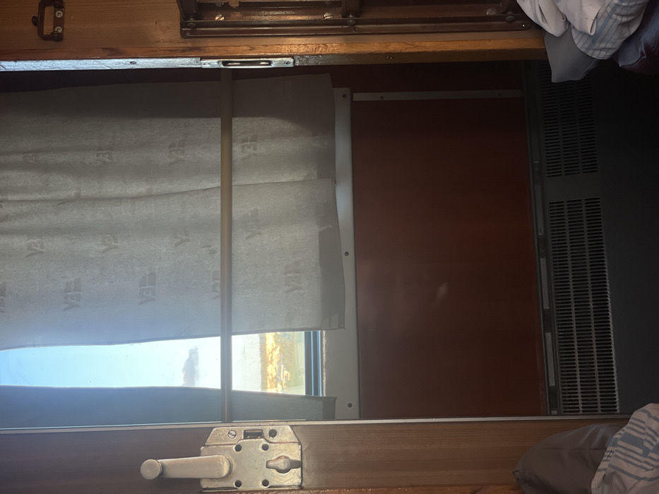 A close-up shot of a train window covered by a slightly raised, patterned curtain. A wooden rod is positioned vertically in front of the curtain. Below the window, a metal latch mechanism is visible. To the right of the window, there's a dark brown, possibly wooden panel and a gray, ventilated section. Above, there's dark brown wooden panelling with some metal fixtures and some light coloured fabric/clothes.