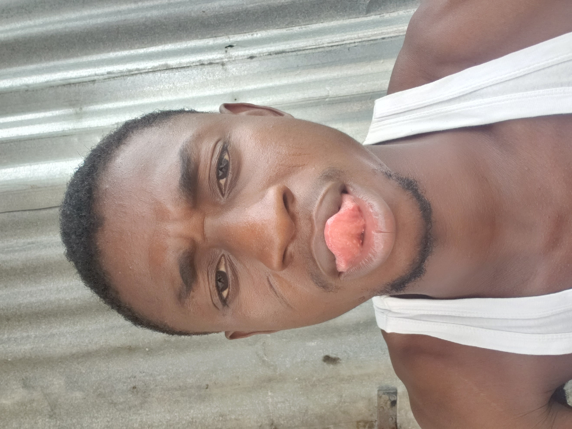 A man with dark skin, short black hair, and a white tank top is photographed outdoors. He is sticking his tongue out and has a slight frown. The background appears to be a weathered metal surface. The location is Adankolo, Nigeria.