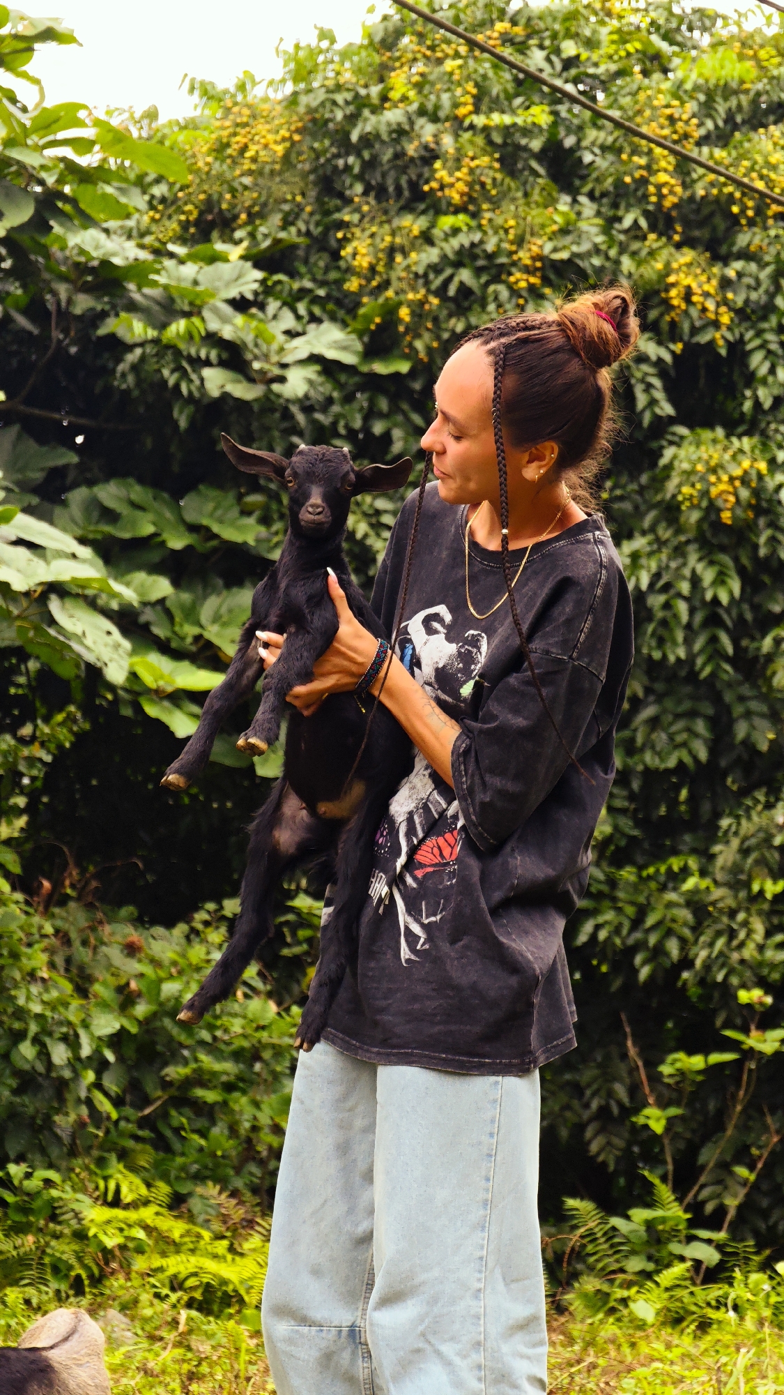 A young Black goat is held by a person wearing a dark graphic t-shirt and light blue wide-leg jeans. The person has their hair in braids pulled up into a bun and is wearing jewelry, including a gold chain and bracelets. The goat is looking towards the camera, with its ears pointed upwards. The background consists of lush green foliage with yellow flowers, suggesting an outdoor, possibly rural, environment. The lighting indicates it is daytime.