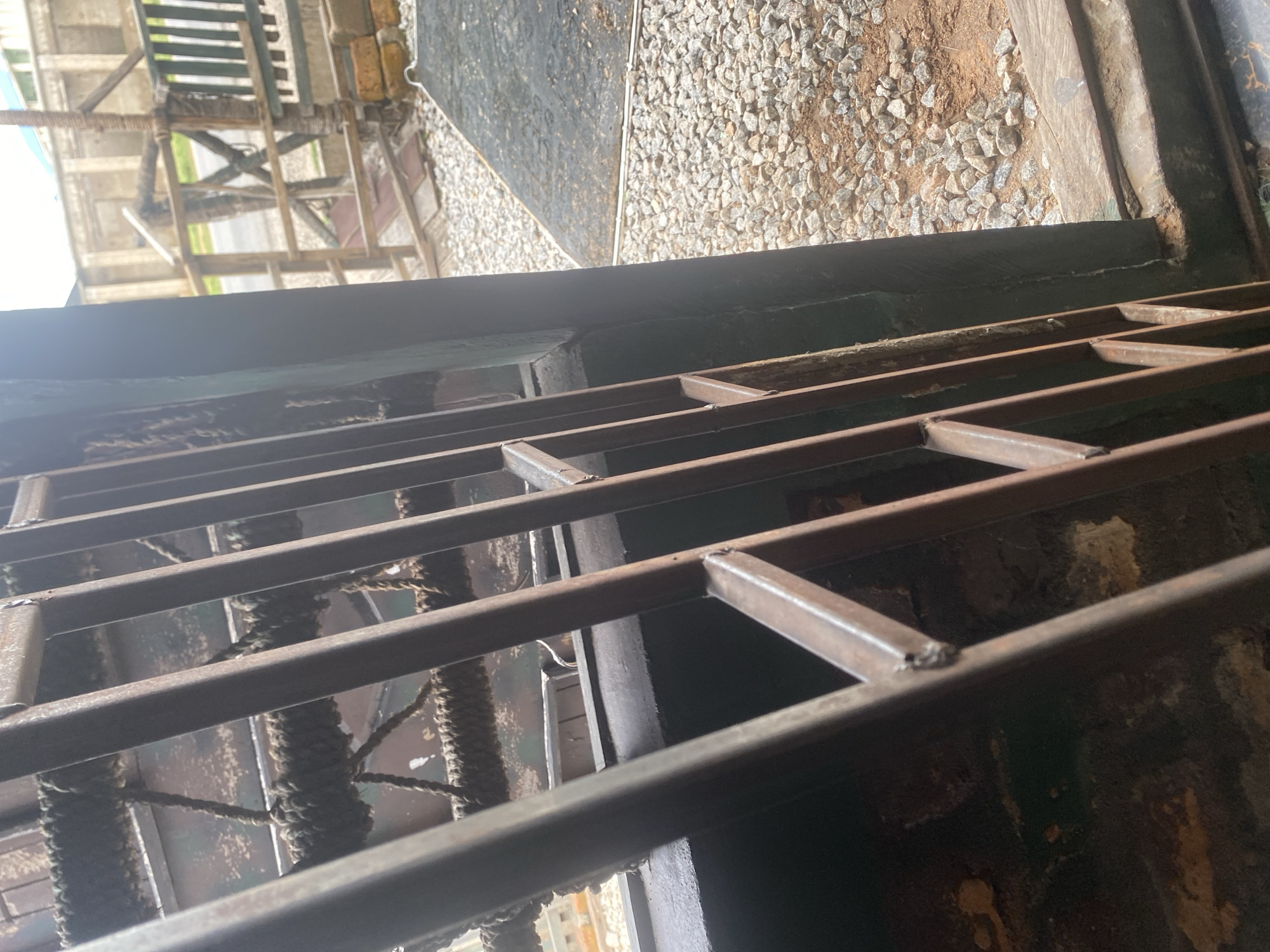 This image captures a close-up view of a metal grate, likely part of a fence or barrier, viewed from below. The grate is constructed from square metal tubing, welded together to form a series of rectangular openings. Through the openings, thick, weathered ropes are visible, wrapped around vertical supports or possibly used for some form of construction or binding. The ropes appear to be made of natural fibers and show signs of wear and age.
Beyond the grate and ropes, the background is blurred but suggests an outdoor setting. There are glimpses of what could be wooden structures, possibly scaffolding or a building under construction, with beams and planks visible. In some areas, a surface covered with small, light-colored stones or gravel is discernible, hinting at an unpaved ground or pathway. The lighting suggests it is daytime, with natural light illuminating the scene, though the overall tone is somewhat dim, possibly due to the angle of the shot or shade cast by the grate itself.
The scene appears utilitarian and industrial, lacking any immediate signs of human presence, though the ropes suggest recent or ongoing activity. The location context of Jalingo, Nigeria, coupled with the rustic materials and potential construction elements, could point to a site undergoing development or maintenance in a more rural or semi-urban environment. There is no visible text in the image.