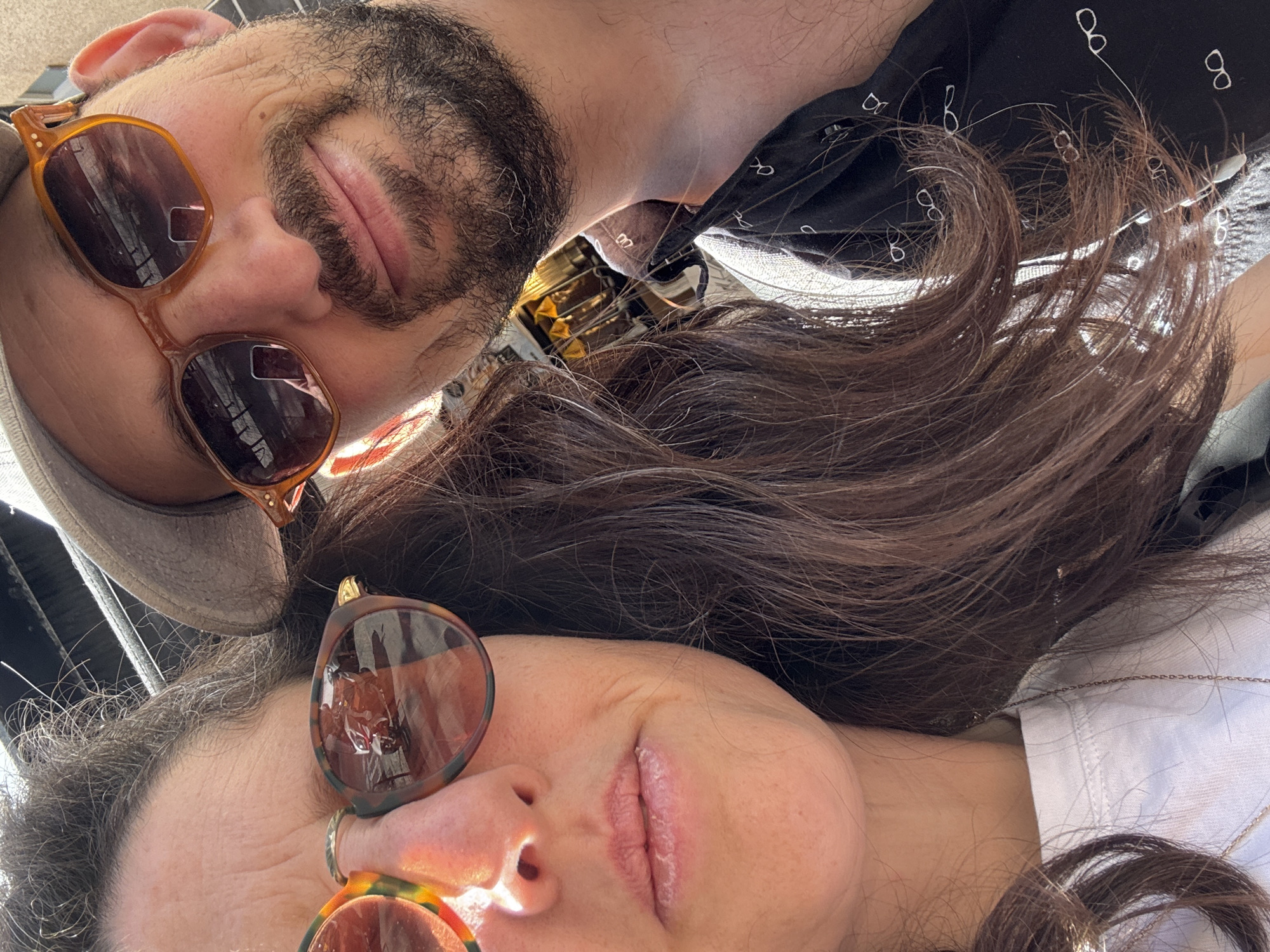 A close-up, upward-angled selfie captures two individuals, likely a couple, in a relaxed and intimate pose. The man is positioned on the left side of the frame, wearing a light-colored cap and amber-toned, rectangular sunglasses. He has a dark beard and appears to have his eyes closed or squinting behind the shades, his lips slightly parted. He is wearing a dark shirt patterned with small white eyeglasses.
The woman is on the right, with long, dark brown, wavy hair cascading across the frame. She wears tortoiseshell-patterned, round sunglasses and a white top, with a thin chain necklace visible around her neck. Her face is also relaxed, with her lips slightly parted.
The image is brightly lit by natural sunlight, indicating it was taken during the day. Strong glares are visible on the sunglasses, and reflections suggest an overhead structure or open sky. The low camera angle implies the subjects are lying down or leaning back, looking up. The overall mood conveys comfort and closeness. This moment takes place in Tel-Aviv, Israel. The repeating eyeglasses pattern on the man's shirt is a notable visual detail.