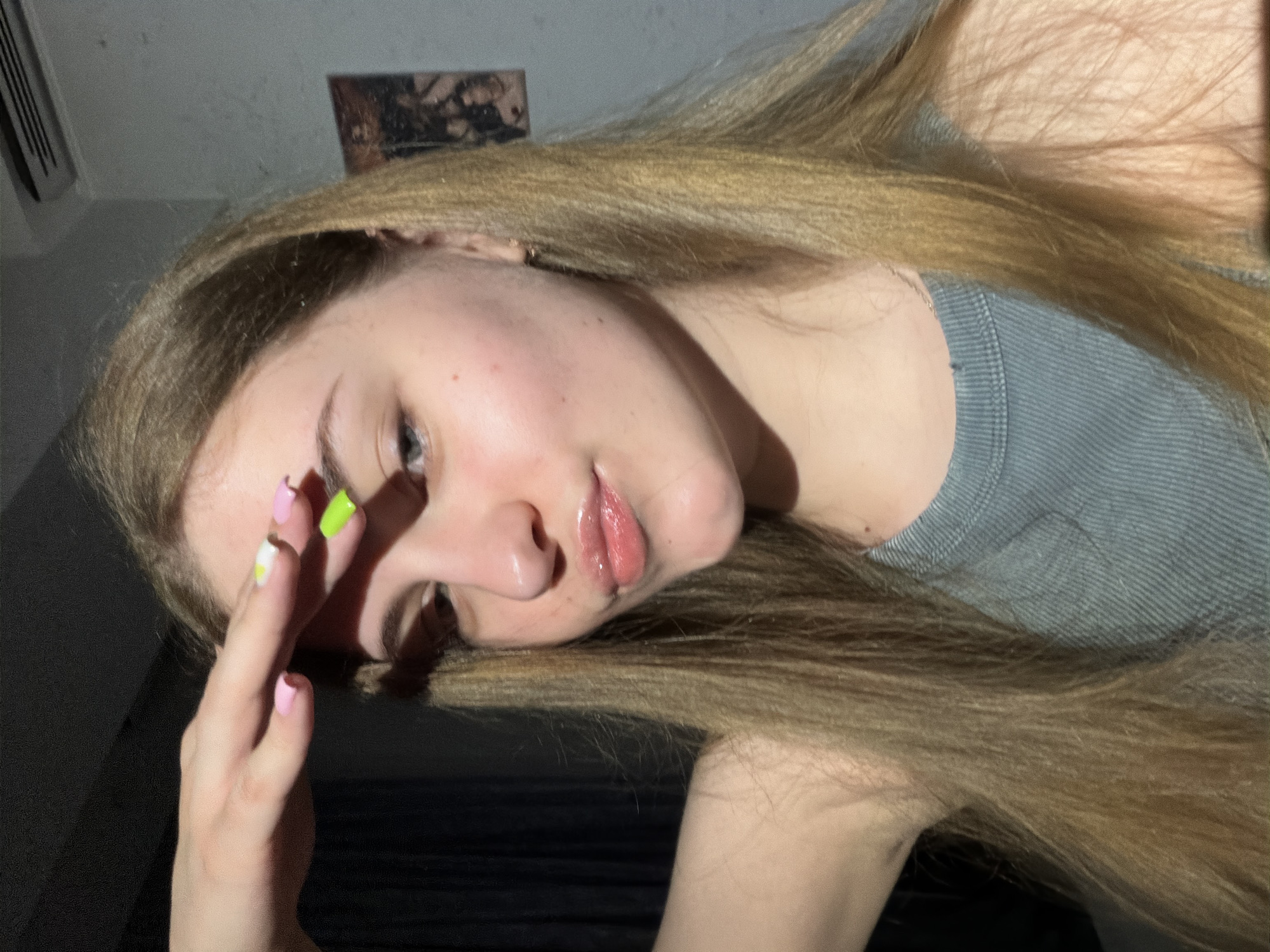 A young woman with long blonde hair is taking a selfie. She is wearing a light blue tank top and has brightly colored nails. Her hand is raised to her forehead, obscuring her eyes. The lighting suggests the sun is shining from the side, creating a shadow across her face. The background appears to be an indoor setting, possibly a bedroom, with a dark curtain on the left and a light-colored wall with some indistinct pictures or decorations on the right. There is an air conditioning unit visible on the wall. The image is focused on the woman's face and upper body. There is no visible information to determine the location in Dnipro, Ukraine.