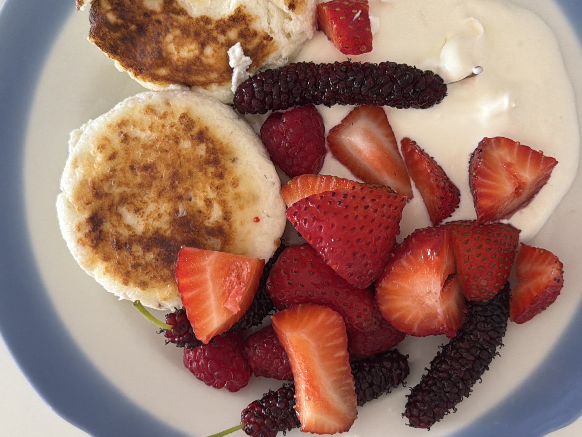 The image shows a plate of food. There are two browned, round items that appear to be pancakes or fritters. The plate also contains a variety of cut strawberries, raspberries, and several elongated dark purple fruits that resemble mulberries. The food is presented on a white plate with a blue rim, and a white sauce or yogurt is also visible. The image provides no information to determine if the image was taken in Tel-Aviv, Israel.