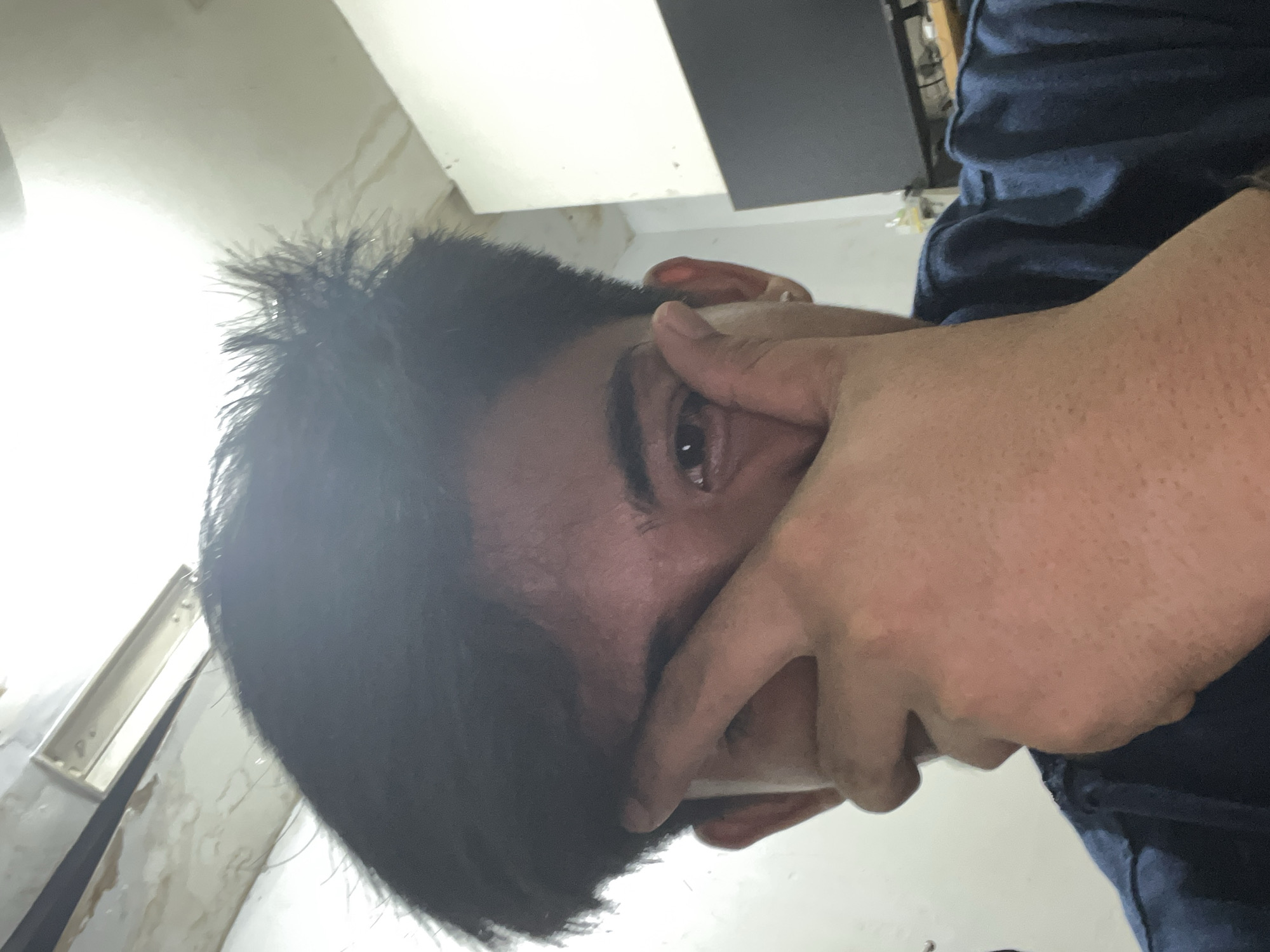 A young person is captured in a close-up, indoor shot, with their right hand covering the majority of their face, obscuring their eyes and nose. Their left eye is visible, appearing brown. They have dark, messy hair styled upwards, and are wearing a dark blue, collared shirt. The background shows a white wall and a light fixture on the ceiling. The image appears to have been taken in a room, possibly a bedroom or living area, located in Central Jakarta, Indonesia.