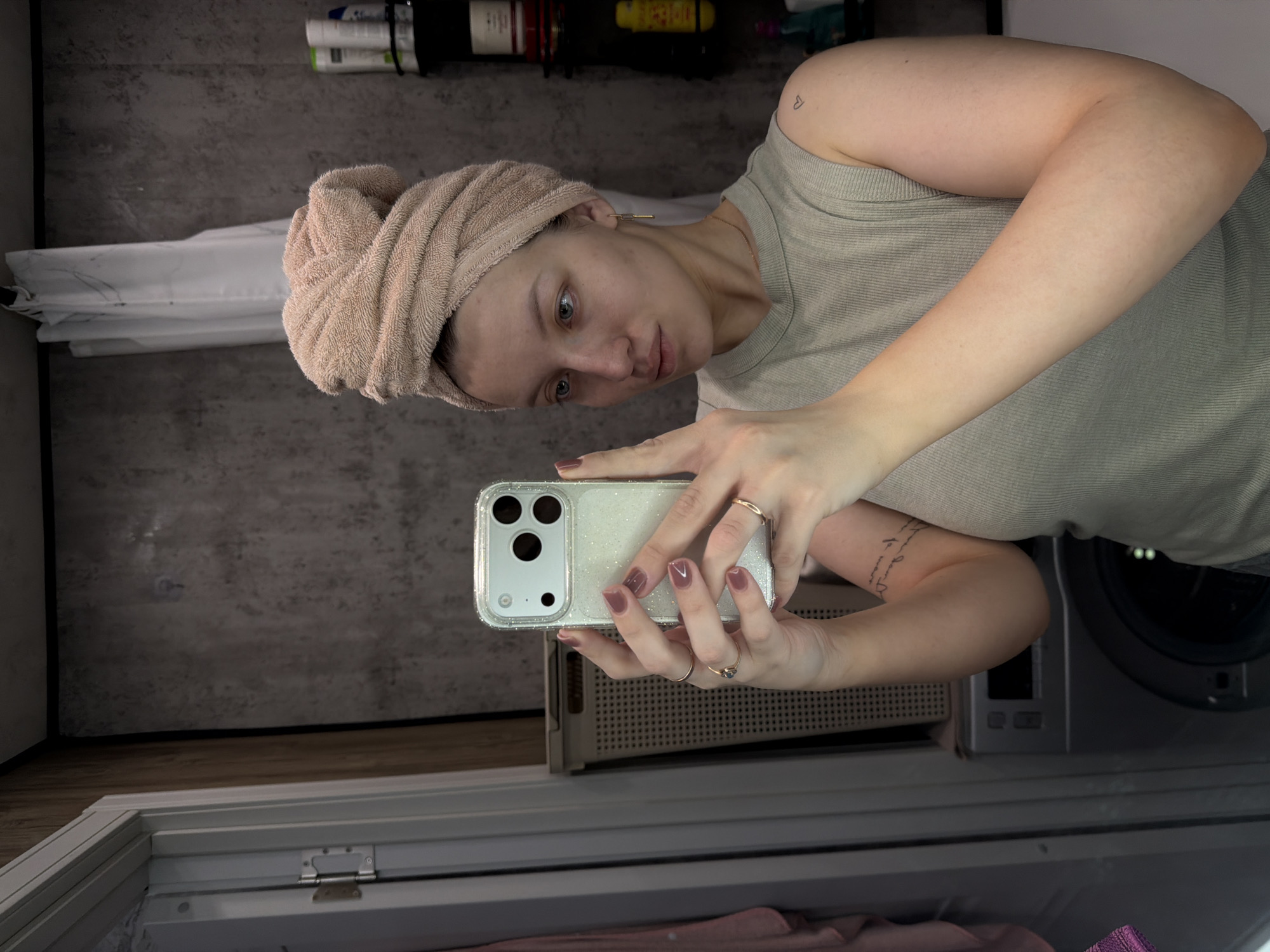A woman with a towel wrapped around her head is taking a selfie in what appears to be a bathroom or laundry room setting. She is holding a smartphone in her hands, and her facial expression suggests she is looking at the screen. The background includes a washing machine, storage shelves with various items, and a mirror reflecting part of the scene. No specific landmarks or details indicate the exact location in Reshetnikovo, Russia.