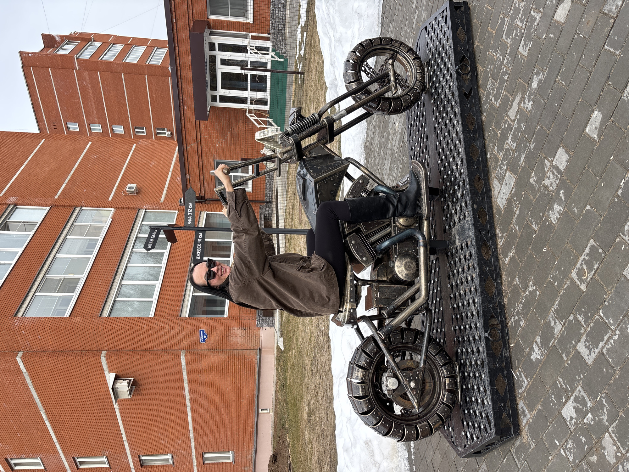 An adult female is depicted seated on a large, dark metal sculpture designed as a custom motorcycle. The sculpture, featuring industrial detailing with exposed pipes, rivets, and chained wheels, is mounted on a rectangular, textured dark metal platform. The woman, smiling and wearing sunglasses, a brown jacket, black pants, and black boots, holds the sculpture's handlebars. The scene is located on Kabanovskaya Street, 24/1, in Chaikovsky, Russia, identified by a blue street sign on the adjacent red-brick, multi-story building. A black directional signpost in the background indicates distances to Moscow (1264 km), Izhevsk (91 km), and Ufa (312 km), confirming the city. Patches of melting snow are visible on the dormant grass bordering the grey brick paved area where the sculpture is situated. Another similar red-brick building is visible further in the background.
