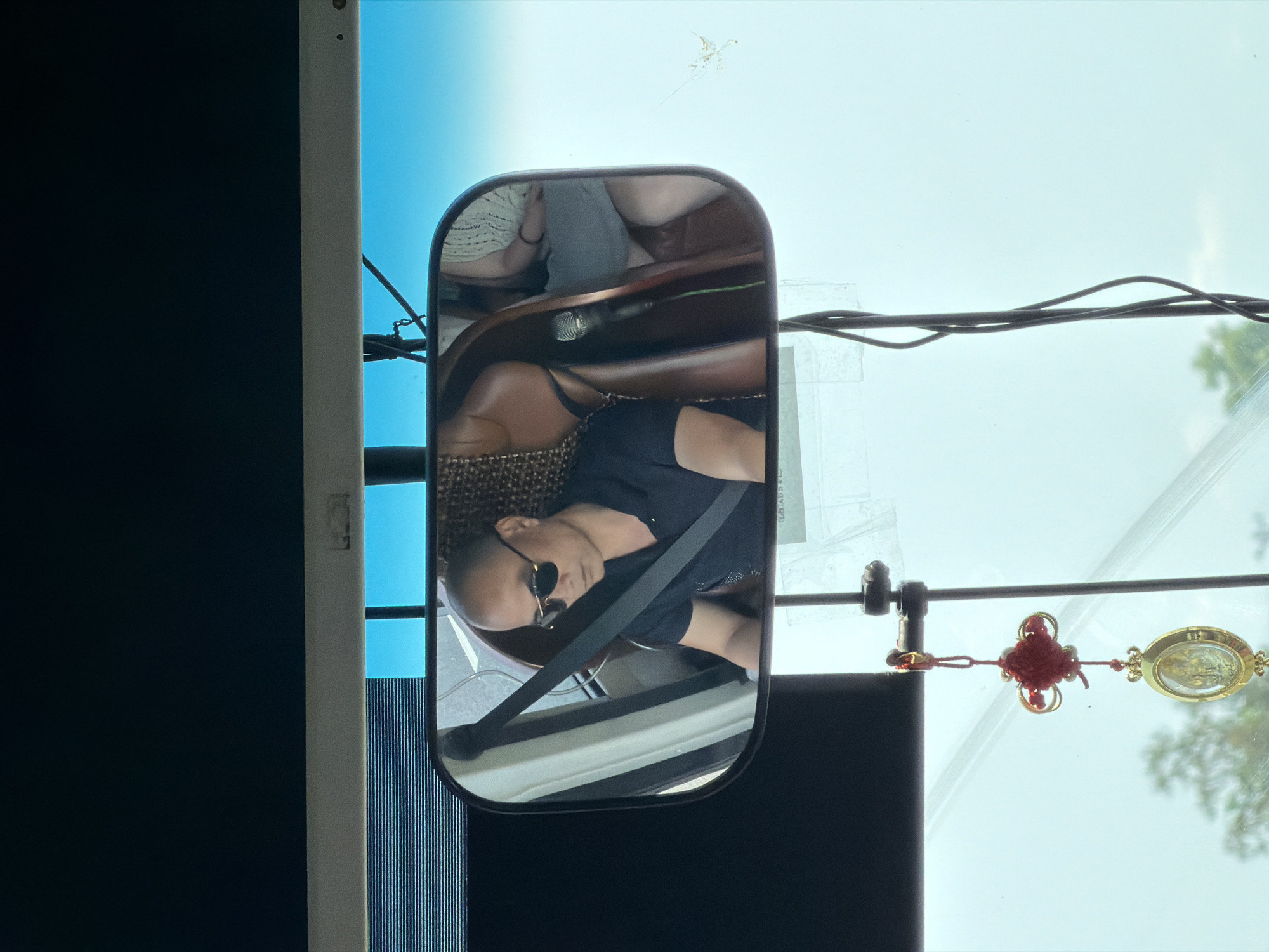 The reflection in a bus side mirror shows a person wearing sunglasses and a seatbelt. A red Chinese knot and a religious pendant hang from the bus's interior near the windshield. Outside the bus, power lines are visible against a hazy sky and some tree branches. The location is likely in Hải Phòng, Vietnam, based on the red Chinese knot and religious pendant which are commonly found in this area of the world.