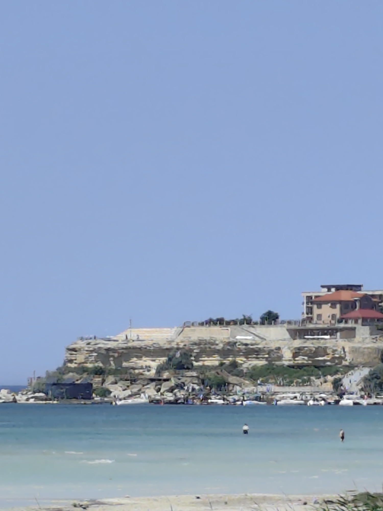This image depicts a coastal scene in Aktau, Kazakhstan, featuring a rocky cliff face overlooking a body of water. On top of the cliff, several buildings are visible, including a multi-story structure with a red-tiled roof and a lighter-colored facade, and another building with a dark roof. The cliff is reinforced in some areas with man-made structures, suggesting development or fortifications. Green vegetation is interspersed with the rocks.
At the base of the cliff, the water is a clear turquoise, transitioning to lighter shades closer to the shore, indicating shallow depths. Several small boats are moored near the shore. Two individuals are visible in the water, wading or swimming. One person is closer to the shore, and another is further out. The sky is a clear, pale blue. The foreground shows a sandy beach area with some dry vegetation. The overall impression is a sunny day at a developed coastal location.