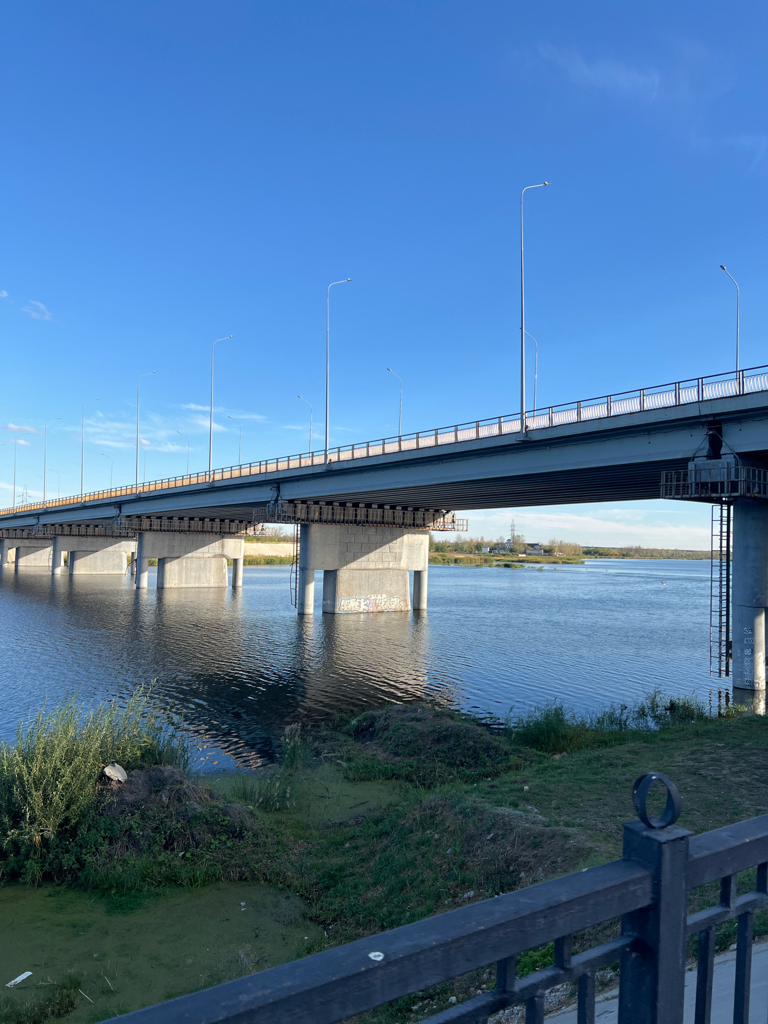 A wide, multi-span concrete bridge stretches across a calm, blue river under a clear, bright sky. The bridge has a gray railing and numerous streetlights. The riverbanks are lined with green grass, some with patches of algae on the water's surface. In the distance, trees and buildings are visible on the far bank. The image captures the bridge from a slightly low angle, emphasizing its scale.