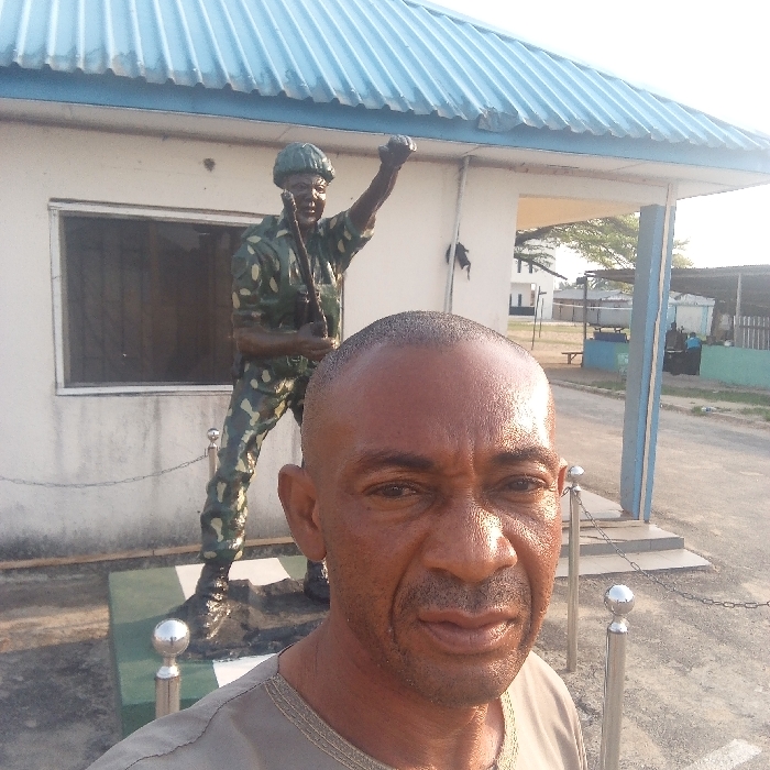 An outdoor daylight photograph in Koko, Nigeria, features an adult male positioned for a selfie in the foreground, his upper torso visible. He has a shaved head, dark skin, and a light goatee, wearing a grey shirt with patterned trim, and looks directly at the camera. Behind him, to the left, stands a life-sized statue of a soldier, painted in green and brown camouflage, holding a rifle in the right hand and with the left fist raised. The statue rests on a rectangular green and white pedestal. Immediately behind the statue is a single-story building with white walls, a barred window, and a light blue corrugated metal roof. To the right, a series of silver bollards connected by chains mark a boundary. The background shows an open area with visible ground, distant trees, and additional structures under a bright sky.
