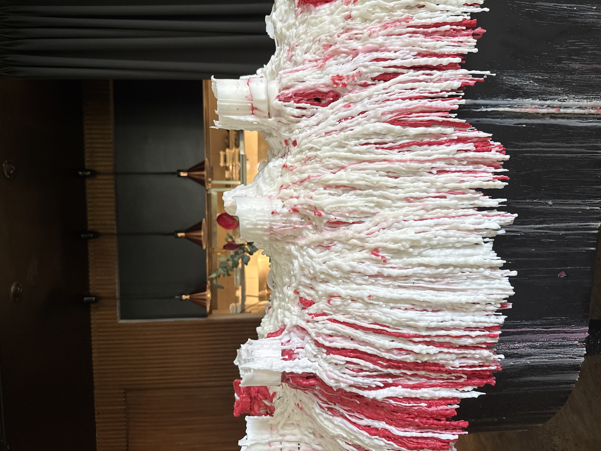 The image displays a close-up of a large, abstract sculpture made from melted white and red wax, resembling dripping candle wax. The sculpture is draped over a dark, cylindrical base, with wax also dripping down the sides of the base. In the background, a wall with vertical wooden slats is visible, along with hanging pendant lights with copper-colored shades. Further back, a dark curtain hangs on the right side, and shelves with books and decorative items are partially visible on the left. The overall impression is that of an artistic installation. The location in Kuta Selatan, Indonesia, is not directly discernible from the visual elements of the photograph.
