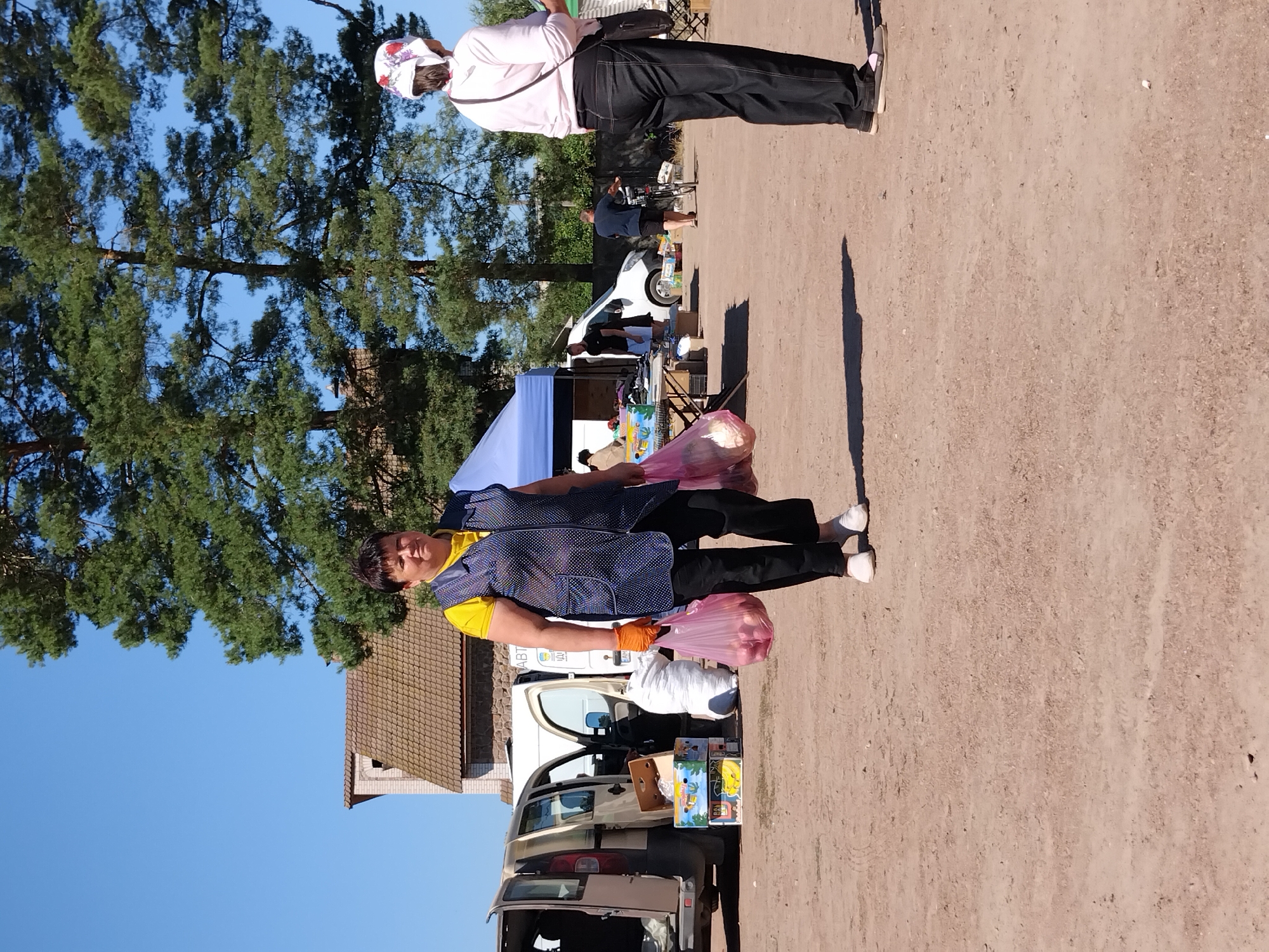 A woman in a blue polka-dot vest over a yellow t-shirt and black pants walks with two pink plastic bags filled with red onions in a dirt lot. She wears orange gloves and white shoes. Behind her, a light-colored van with its rear doors open is parked next to a white cargo van. In the background, a building with a brown-tiled roof stands under tall green trees, and a blue tent is set up. Another woman in a pink shirt and black pants stands facing away from the camera to the right, with a dark bag slung over her shoulder. The scene appears to be a market or an outdoor gathering in Irshansk, Ukraine, under a clear blue sky.
