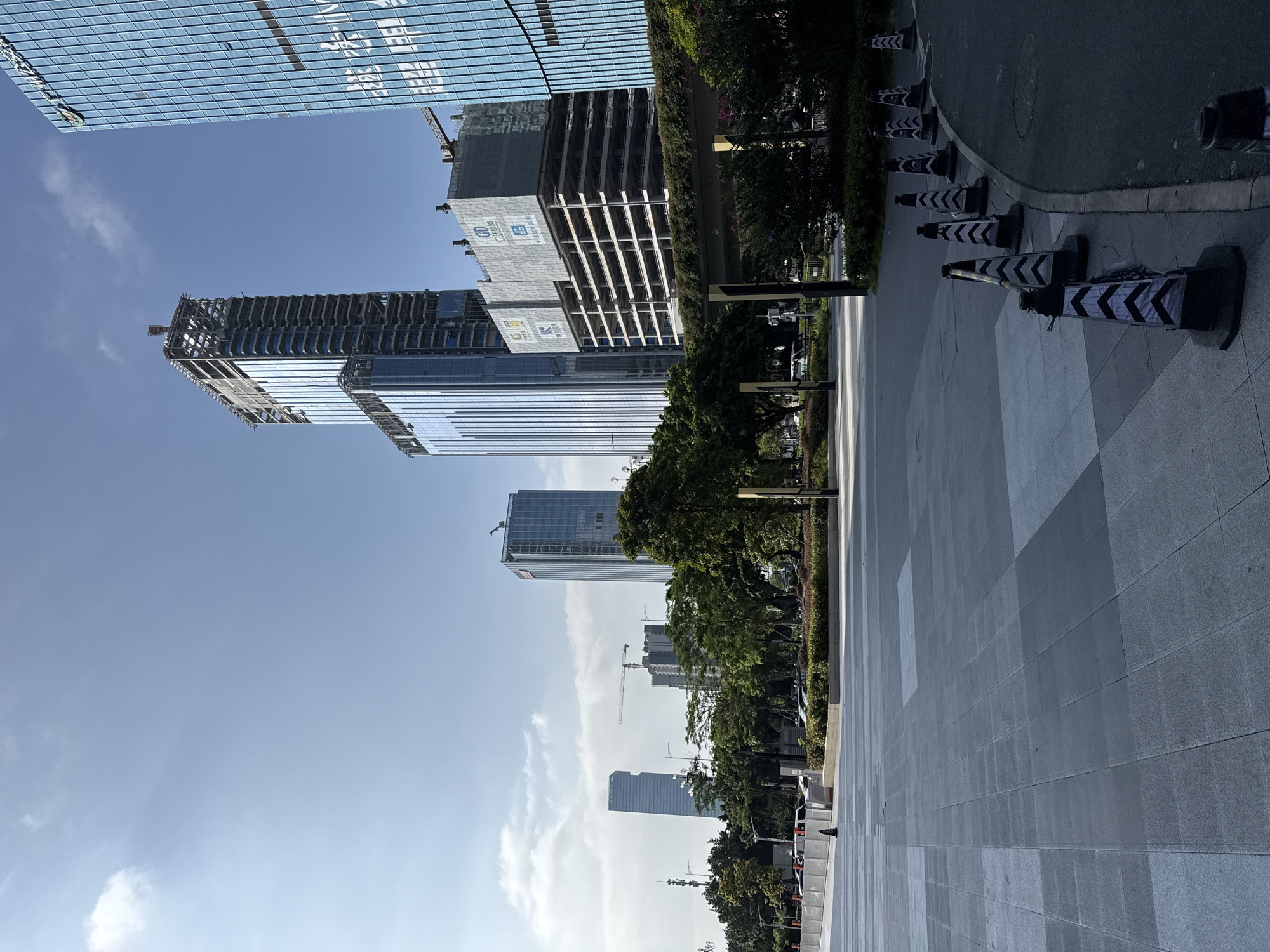 The image depicts a modern city scene in Huangpu District, China, on a clear day. In the foreground, a grey tiled plaza extends forward, with a curved road and a line of black and white traffic cones on the right. Beyond the plaza, a well-maintained green area with trees and shrubs separates the pedestrian space from a raised walkway or bridge.
Dominating the background are several tall buildings. One prominent structure on the right is a modern, glass-fronted skyscraper with visible text on its facade. To its left, a skyscraper is under construction, with its skeletal frame exposed at the top. Further in the distance, other skyscrapers of varying designs and heights are visible, some appearing completed and others potentially under construction, with cranes visible in the background. The sky is a clear blue with a few scattered white clouds. The overall impression is one of urban development and a bustling commercial or business district. No people are visible in the frame.