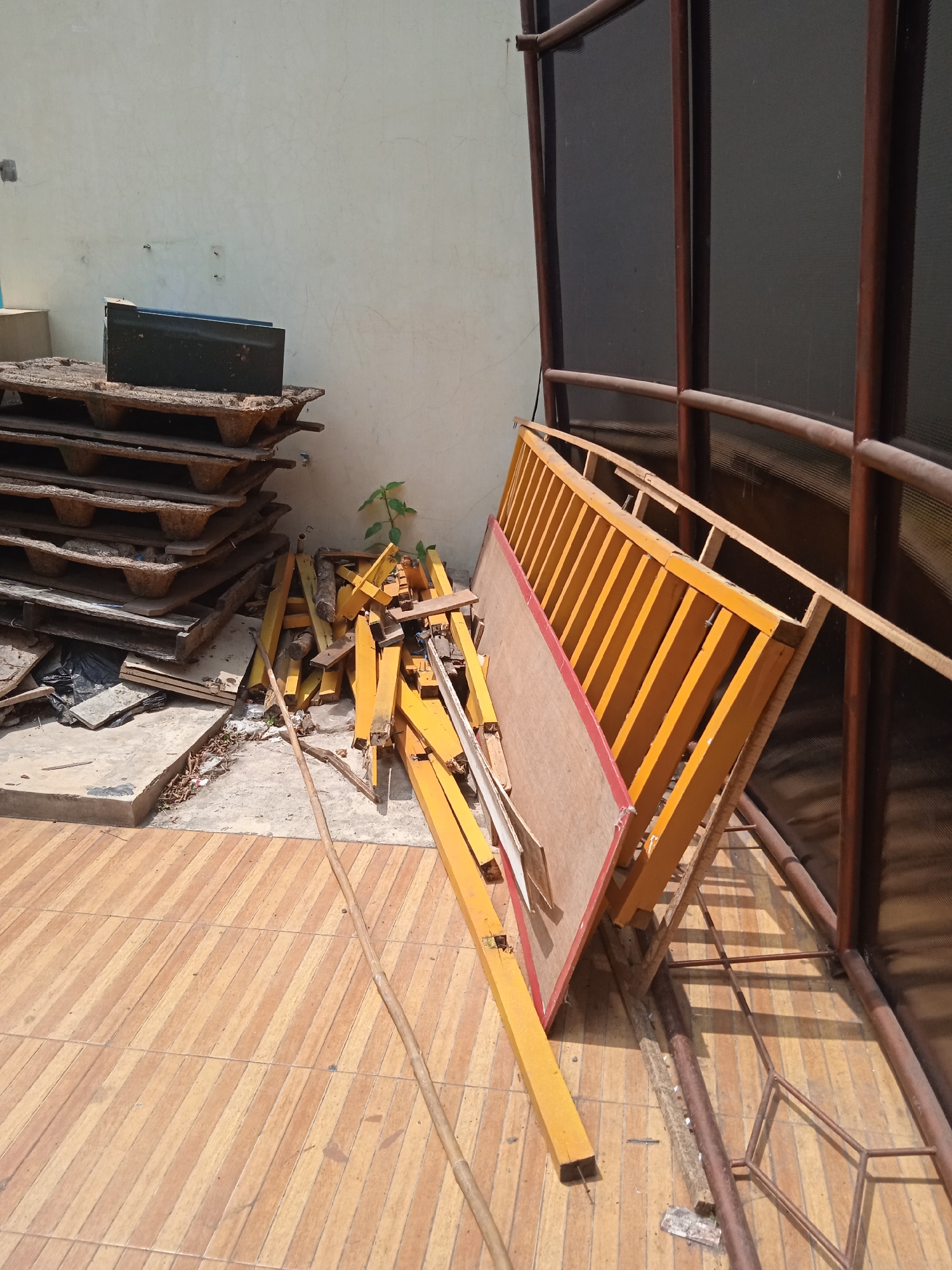 In Cileunyi, Indonesia, an outdoor scene depicts a collection of discarded construction materials and debris. The foreground is dominated by a tiled floor with a diagonal striped pattern in shades of light brown and tan. A long, thin, light brown stick or pole lies diagonally across the tiles, originating from the mid-left and extending towards the bottom right.
To the left, a stack of wooden pallets, weathered and aged, is piled against a pale, textured wall. A dark green metal box rests on top of the uppermost pallet. Between the pallets and the wall, a small, green seedling is visible. Scattered on the ground around the pallets are various pieces of debris, including some black plastic sheeting and broken wooden fragments.
In the center and right of the image, a chaotic pile of bright yellow metal framing and wooden planks is present. Some of these pieces are angled precariously, suggesting they are part of a dismantled structure. A piece of plywood with a reddish-pink border is leaning against the yellow framing.
The background features a portion of a building with a wall and a dark, possibly screened, structure with vertical brown metal supports. The lighting suggests it is daytime, with shadows cast by the objects. The overall impression is one of disarray and abandonment.