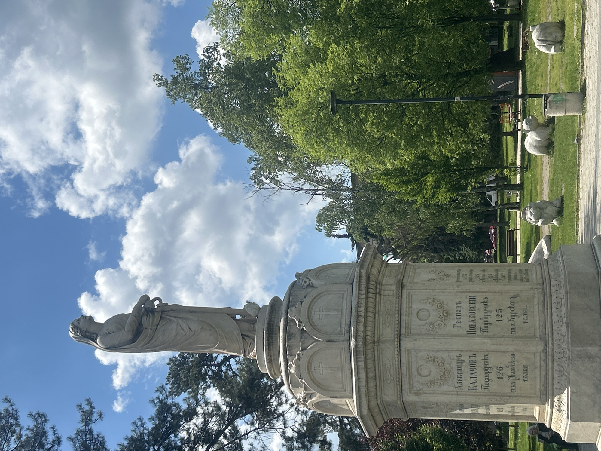 The image shows a marble monument topped with a statue of a reclining figure, possibly in a park or public space in Pleven, Bulgaria. The base of the monument has inscriptions in Cyrillic. The sky is visible with scattered clouds, and trees are in the background. In the distance, there are concrete animal sculptures.