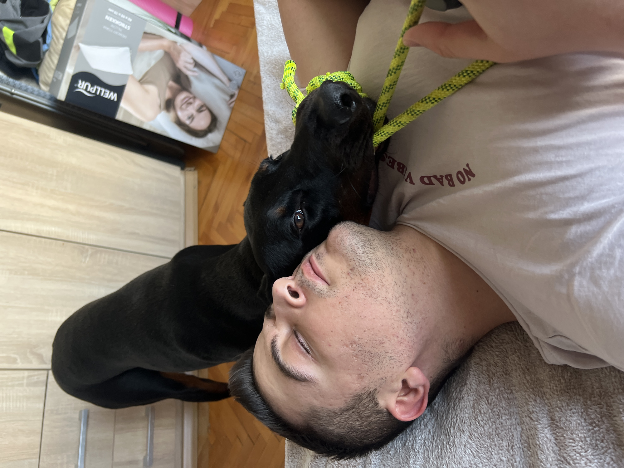 A man is lying on his back on a gray blanket, with a black dog standing over him. The dog's head is near the man's face, and the dog is wearing a neon green rope around its neck. The man's eyes are closed. He is wearing a light-colored t-shirt with the words "NO BAD VIBES" on it. Part of a cardboard box with the word "WELLPUR" is visible in the background, resting on a cabinet with a wooden finish. The floor appears to be made of wood. The location is unspecified, and nothing in the image indicates it is in Sofia, Bulgaria.