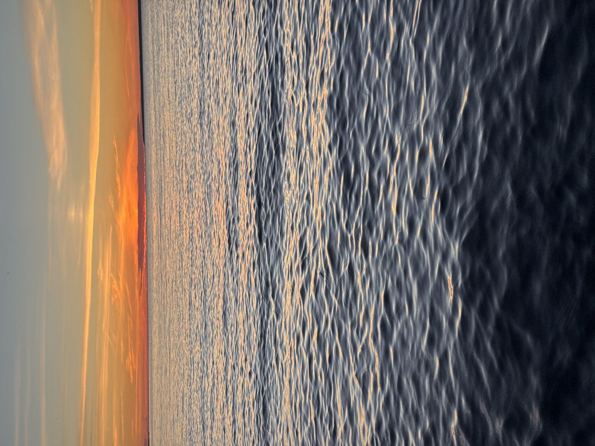 The image depicts a serene seascape at sunset. The sky is a gradient of warm hues, transitioning from a pale blue at the top to vibrant oranges and reds near the horizon. Thin, wispy clouds are scattered across the sky, catching the last light of the day. The sun is setting over a distant landmass, visible as a dark silhouette against the colorful sky. The water is calm, with gentle ripples reflecting the colors of the sky. The foreground is dominated by the dark, textured surface of the water, with highlights of orange and yellow from the sunset scattered across it. There are no people, objects, or actions visible in the image. The scene evokes a sense of tranquility and natural beauty. The specific location in Tallinn, Estonia, is not identifiable from the image alone.