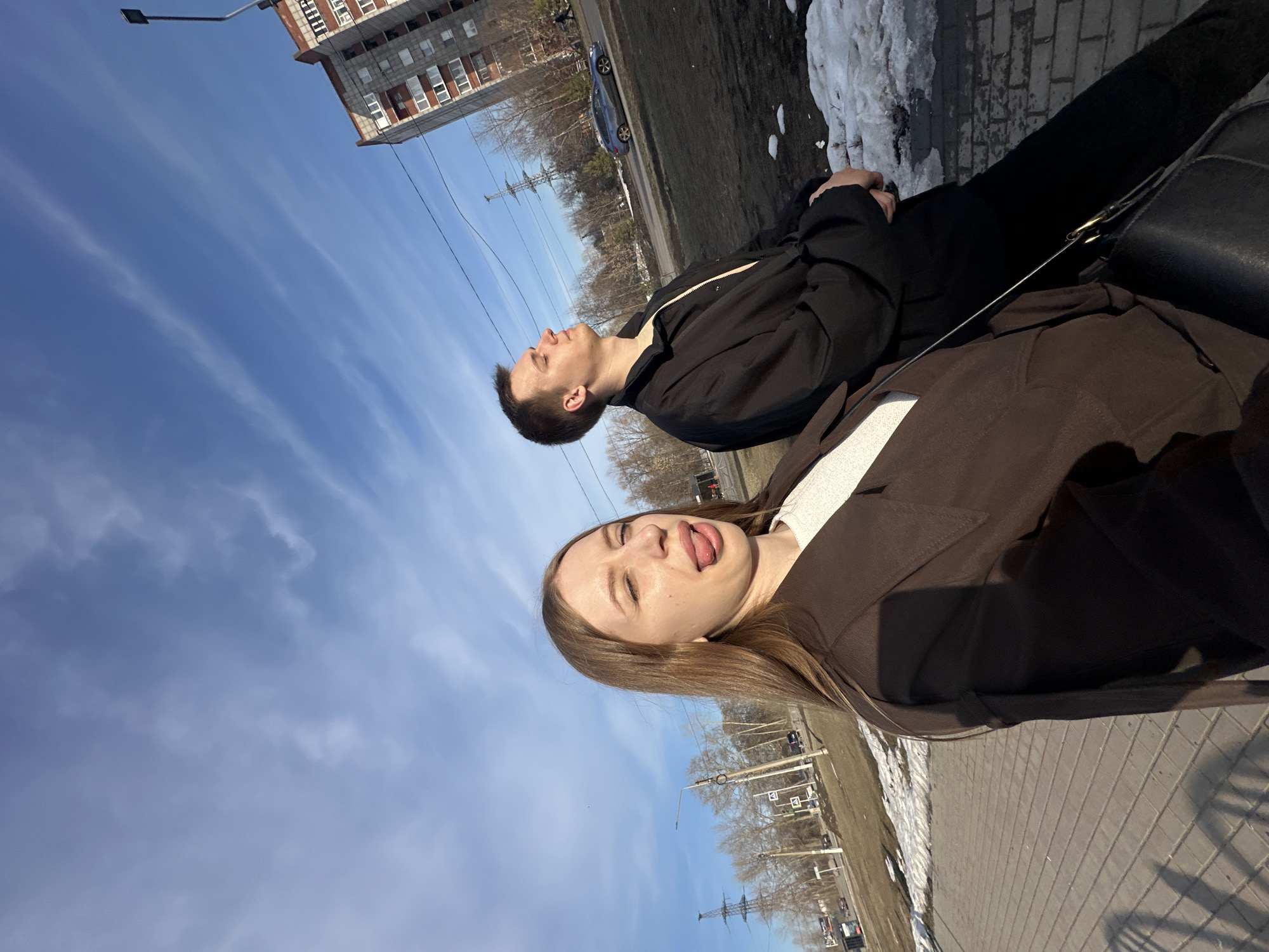 Two people are sitting on a bench outside in Chaikovsky, Russia on a sunny day with a blue, cloudy sky. A woman with light brown hair, wearing a brown jacket over a white shirt, is in the foreground with her tongue sticking out. Behind her is a man wearing a black jacket looking upwards. Behind them are trees, an electrical pole, and a brick building. Next to the bench is grass and a patch of snow. The bench sits on a paved sidewalk.