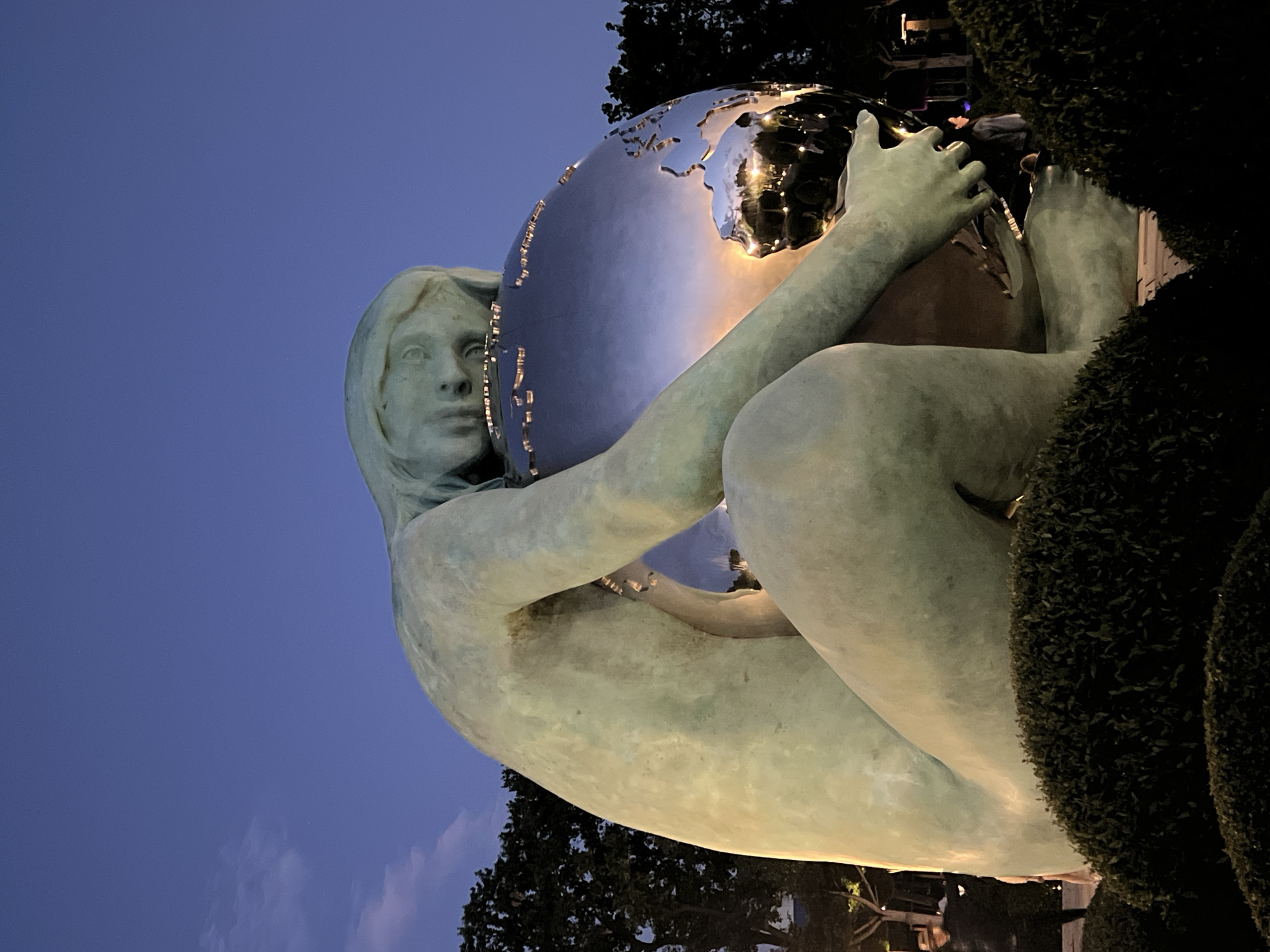 The image depicts "The Embrace of the World" sculpture by Zurab Tsereteli, located in Krasnodar, Russia. The sculpture features a large, nude female figure made of bronze, cradling a mirrored globe. The globe reflects the surrounding environment, including lights and trees. The sculpture is situated in a public space, with manicured bushes in the foreground and trees in the background. The sky is a deep blue, indicating it is either dusk or dawn. A few people are visible in the background, appearing to be pedestrians. The overall impression is one of peaceful contemplation and connection with the world.