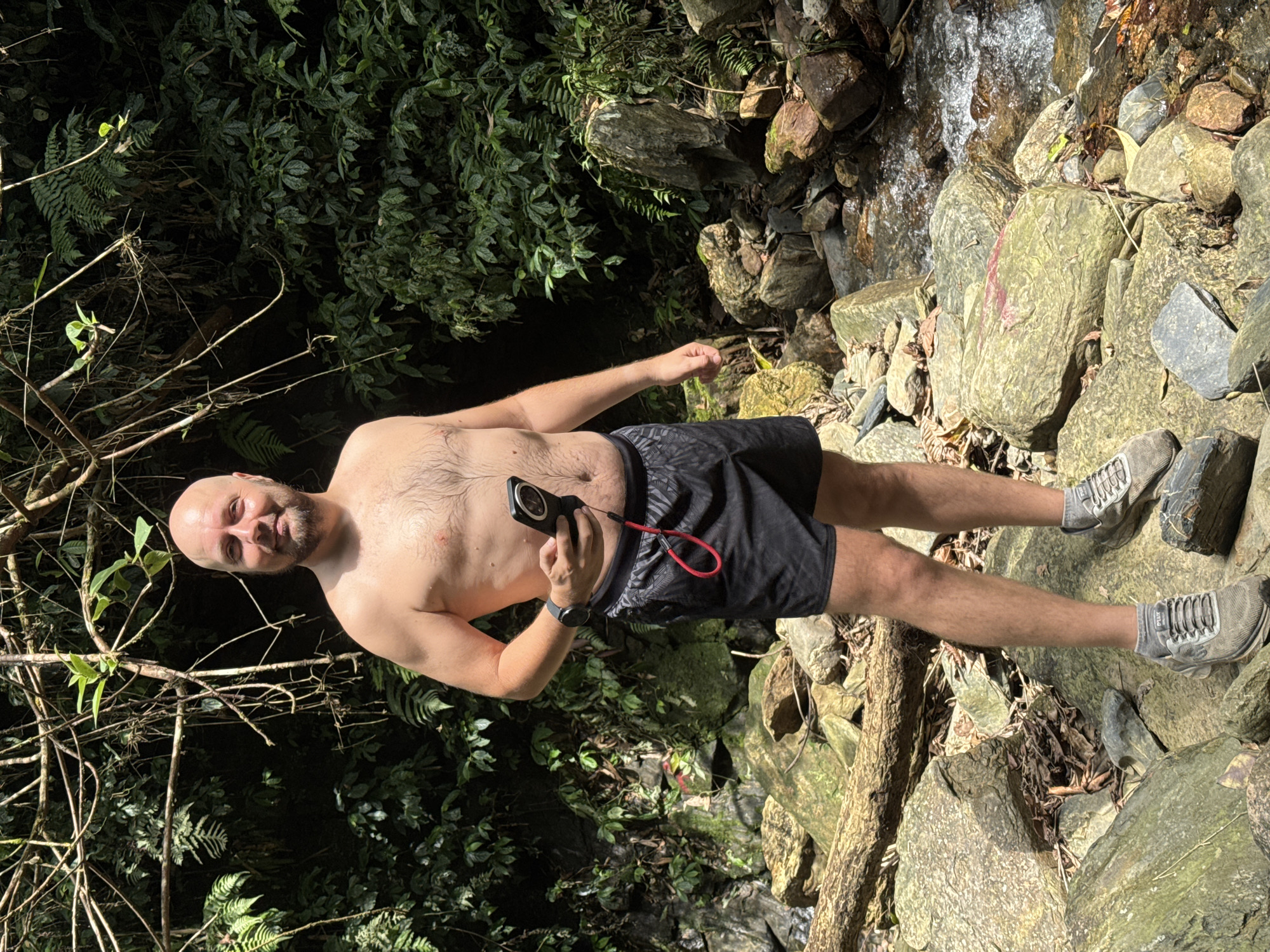 A bald man in black shorts and grey sneakers stands among rocks and foliage, holding a black device with a red cord in his hand. He is shirtless and appears to be in an outdoor environment near a stream or waterfall. The backdrop features lush green vegetation and rocky terrain, suggesting a natural setting. The image was taken in Suối Giàng, Vietnam.