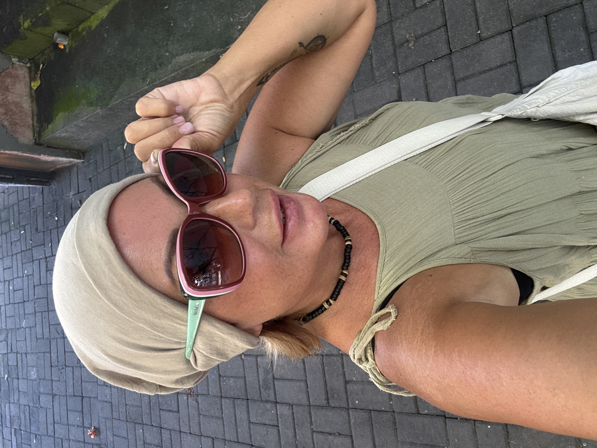 A light-skinned woman is posing for a selfie in Kuta, Indonesia. She is wearing a khaki-colored head wrap, a sleeveless olive green dress, and sunglasses with pink frames and pale green arms. She has a white purse strap across her body, and a black and brown beaded necklace around her neck. Her hair is blonde, and she has a tattoo on her left forearm. The background consists of grey brick pavement. Moss is growing on a concrete edge in the upper left corner of the photo.
