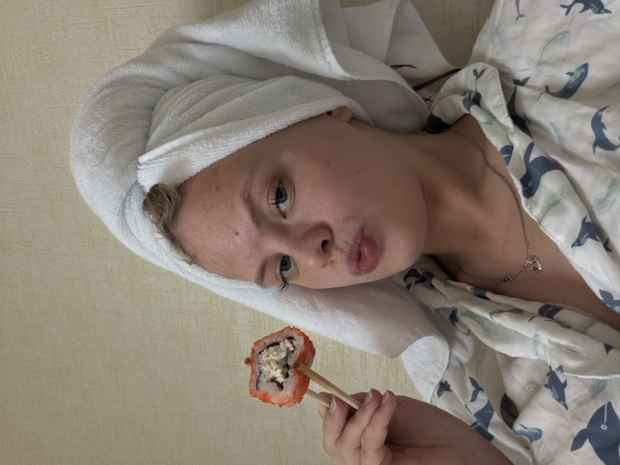 A young woman with a white towel wrapped around her head is lying down, wearing a white shirt with a blue whale pattern, and holding a piece of sushi with chopsticks. She has fair skin, light eyes, and is making a kissing face. A silver necklace with a heart-shaped pendant is visible around her neck. The background is a plain, light-colored wall. The location is not possible to determine from the image.