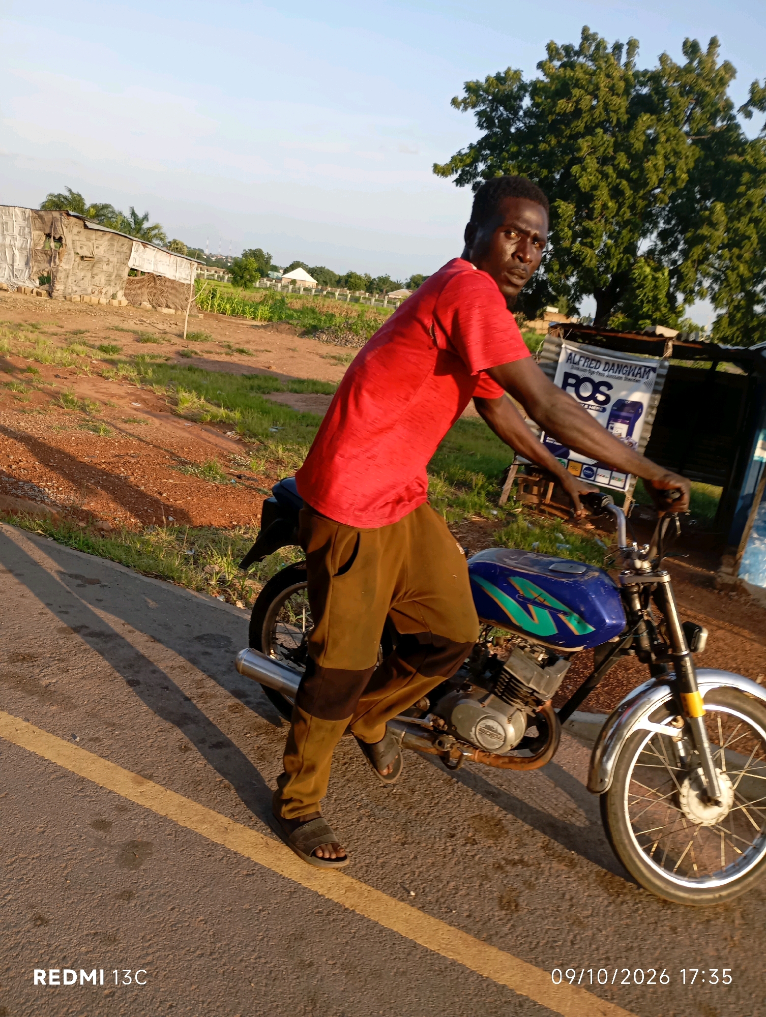 A Nigerian man in a red t-shirt and brown pants is sitting on a blue and green motorcycle. He is looking at the camera with a serious expression. The motorcycle is parked on a road with a yellow line. In the background, there are trees, a building, and a sign that says "ALFRED DANGWHA POS". The image appears to have been taken in Shendam, Nigeria.