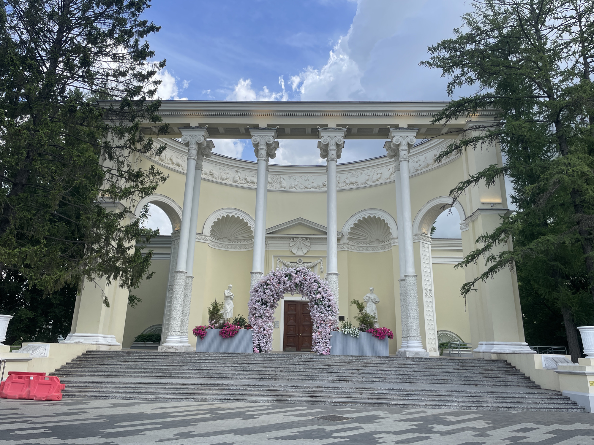This image depicts the Rotunda at VDNKh (Exhibition of Achievements of National Economy) in Moscow, Russia. The neoclassical rotunda features a semi-circular facade with a colonnade of white Ionic columns supporting a decorative entablature. A wide, symmetrical staircase leads up to the entrance, which is adorned with a large floral archway composed of pink and white flowers. Two classical statues stand on either side of the entrance, flanking the floral arch. Planters filled with pink flowers are placed in front of the statues. The building itself is painted a pale yellow and has ornate relief decorations above the arched doorways. Tall evergreen trees frame the structure, and the sky above is a mix of blue and white clouds. In the foreground, a grey patterned pavement is visible, with a red construction barrier on the left side of the steps. The overall scene suggests a festive or ceremonial occasion.